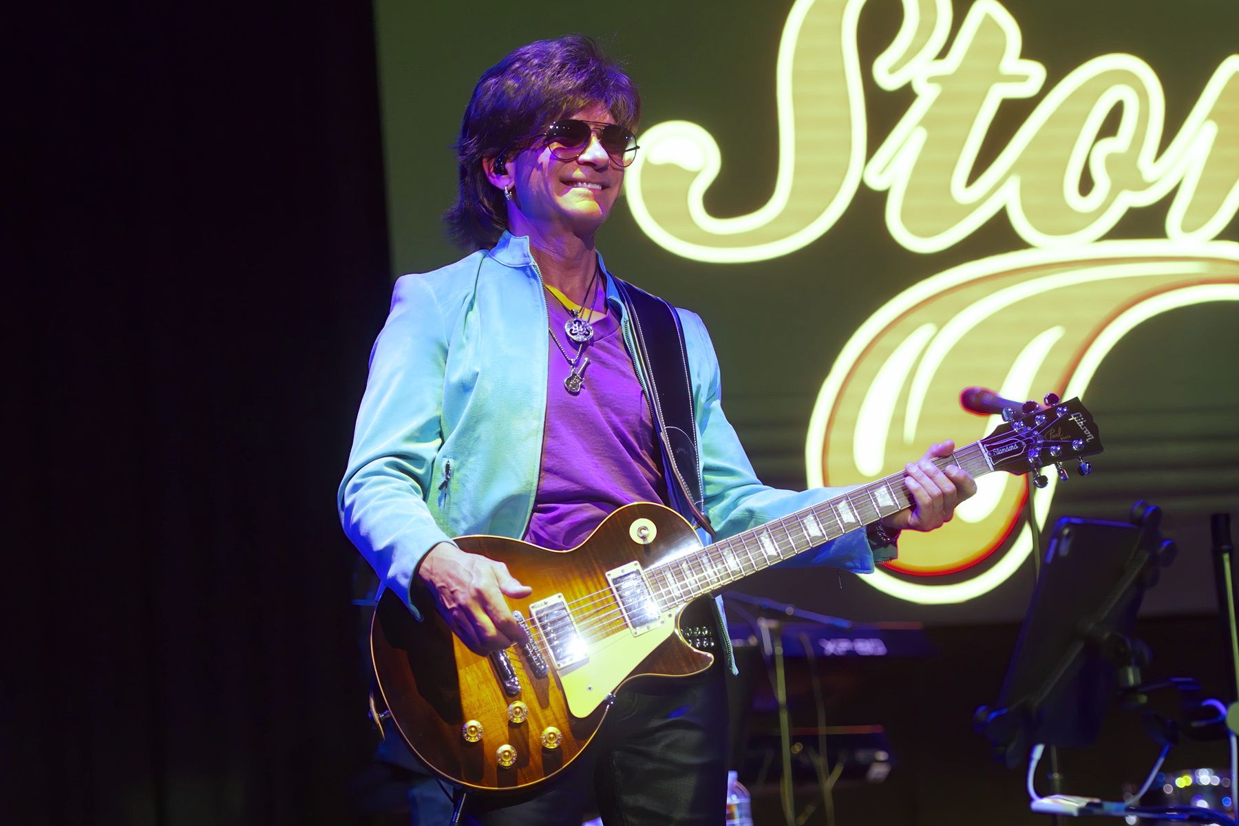 Musician playing electric guitar onstage under purple and green lights, with a neon sign in the background