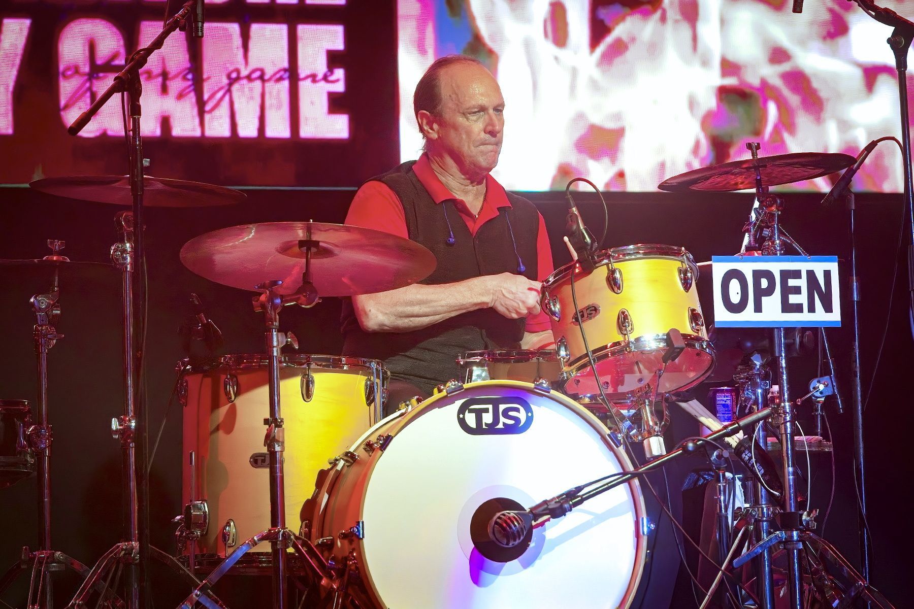 Drummer playing on a stage with a yellow drum kit and red lighting, “OPEN” sign visible nearby