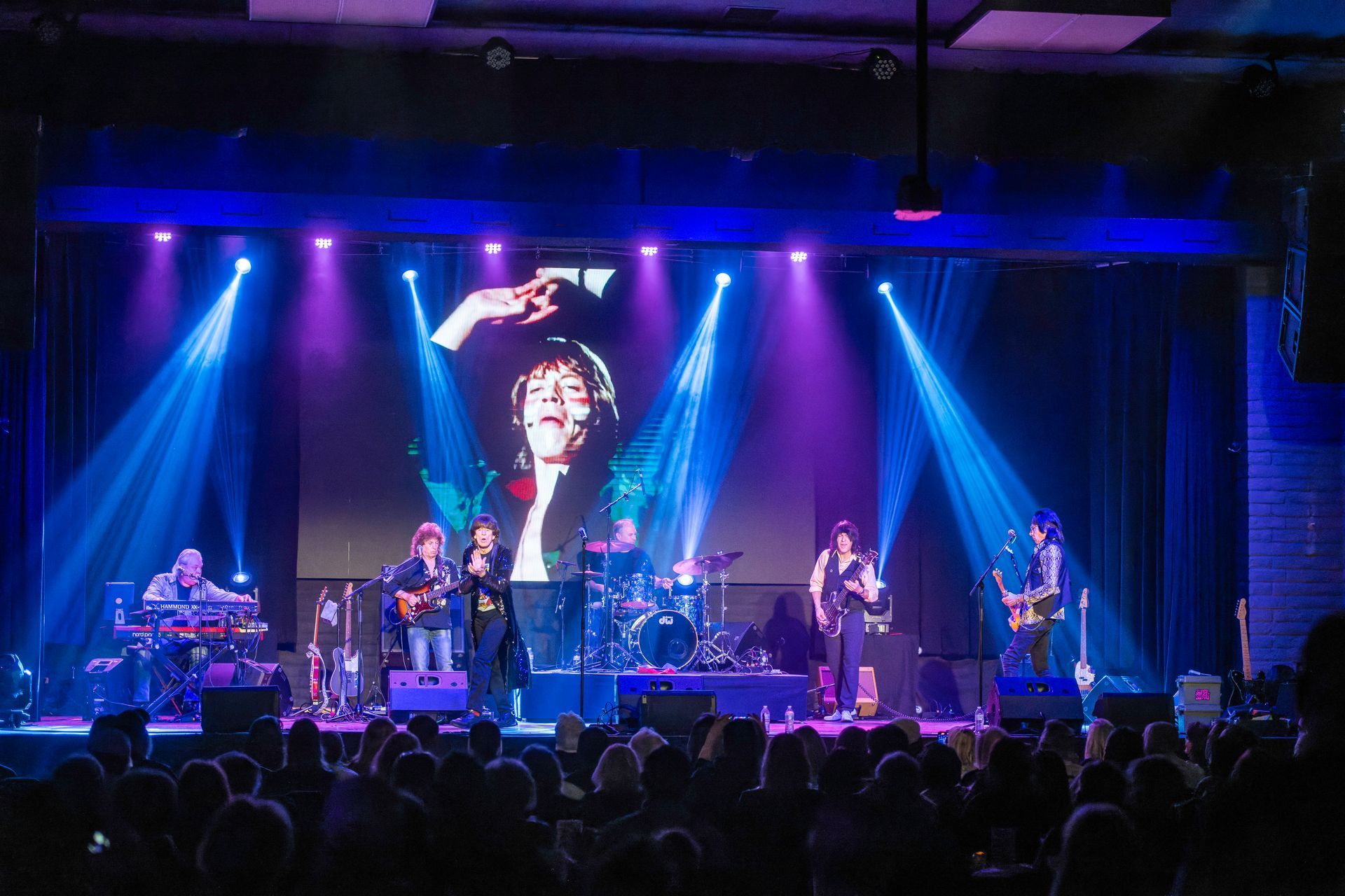 Live concert with blue and purple stage lights, band performing, and a large portrait on the screen behind them