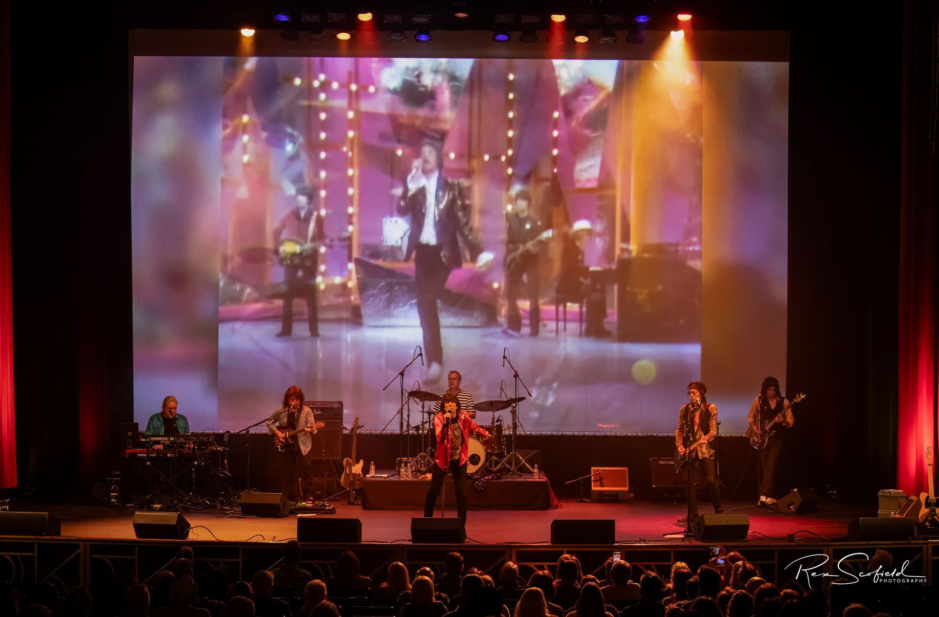 Live band performing on a stage under colorful lights with a large projected backdrop.