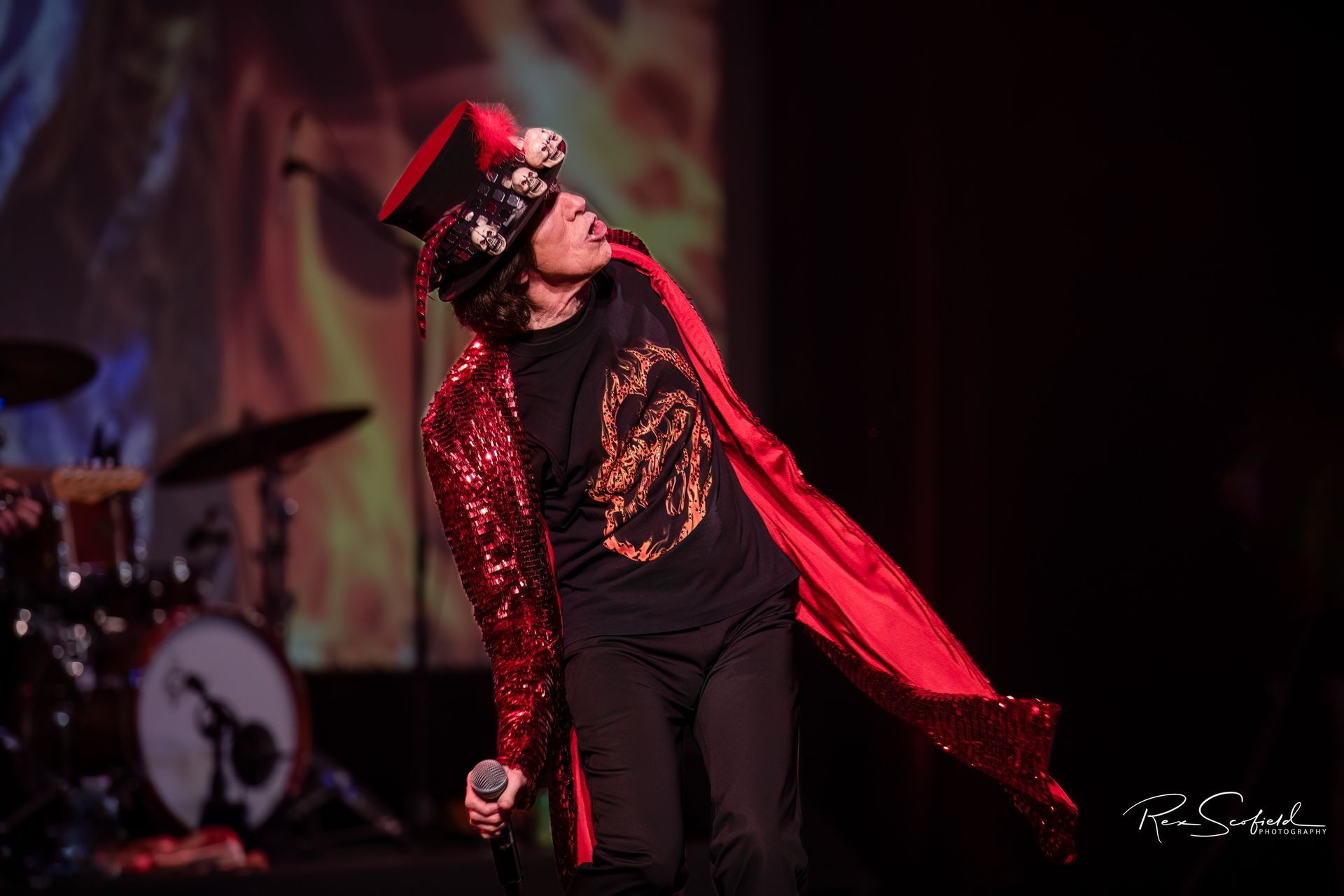 Singer in red coat and black hat performing onstage under red lights