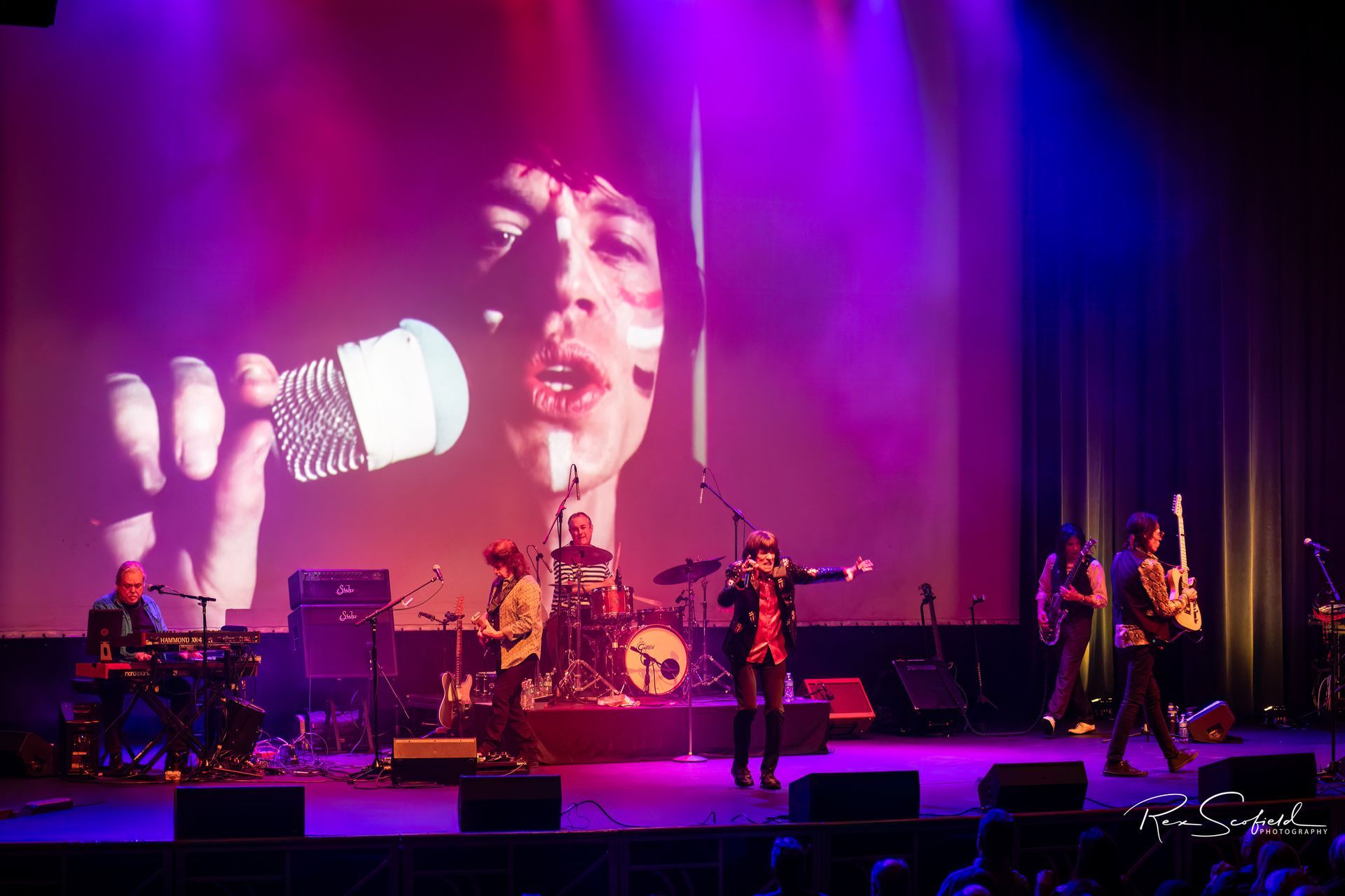 Rock band performing on a purple-lit stage, with a singer projected large on the screen behind them.