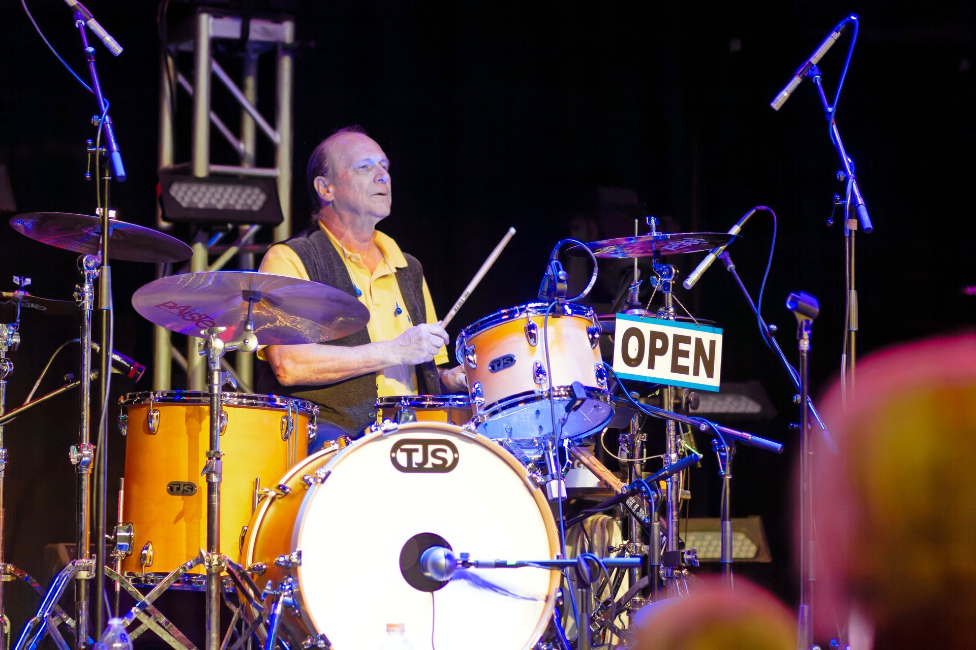 Drummer at a yellow drum kit onstage, playing under purple lights with an “OPEN” sign on the bass drum