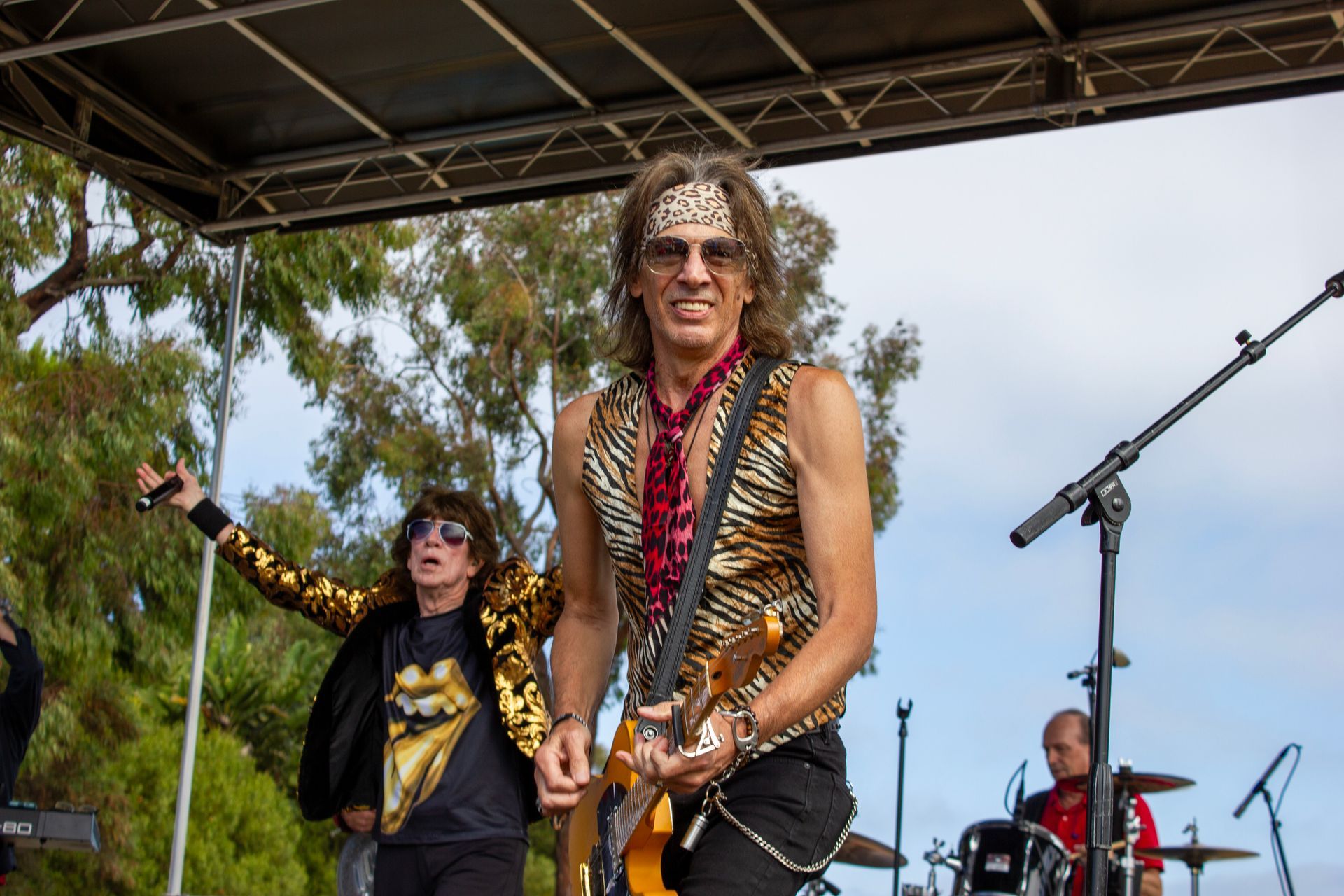 Rock band performing on an outdoor stage, with a guitarist in the foreground and drummer in the background.