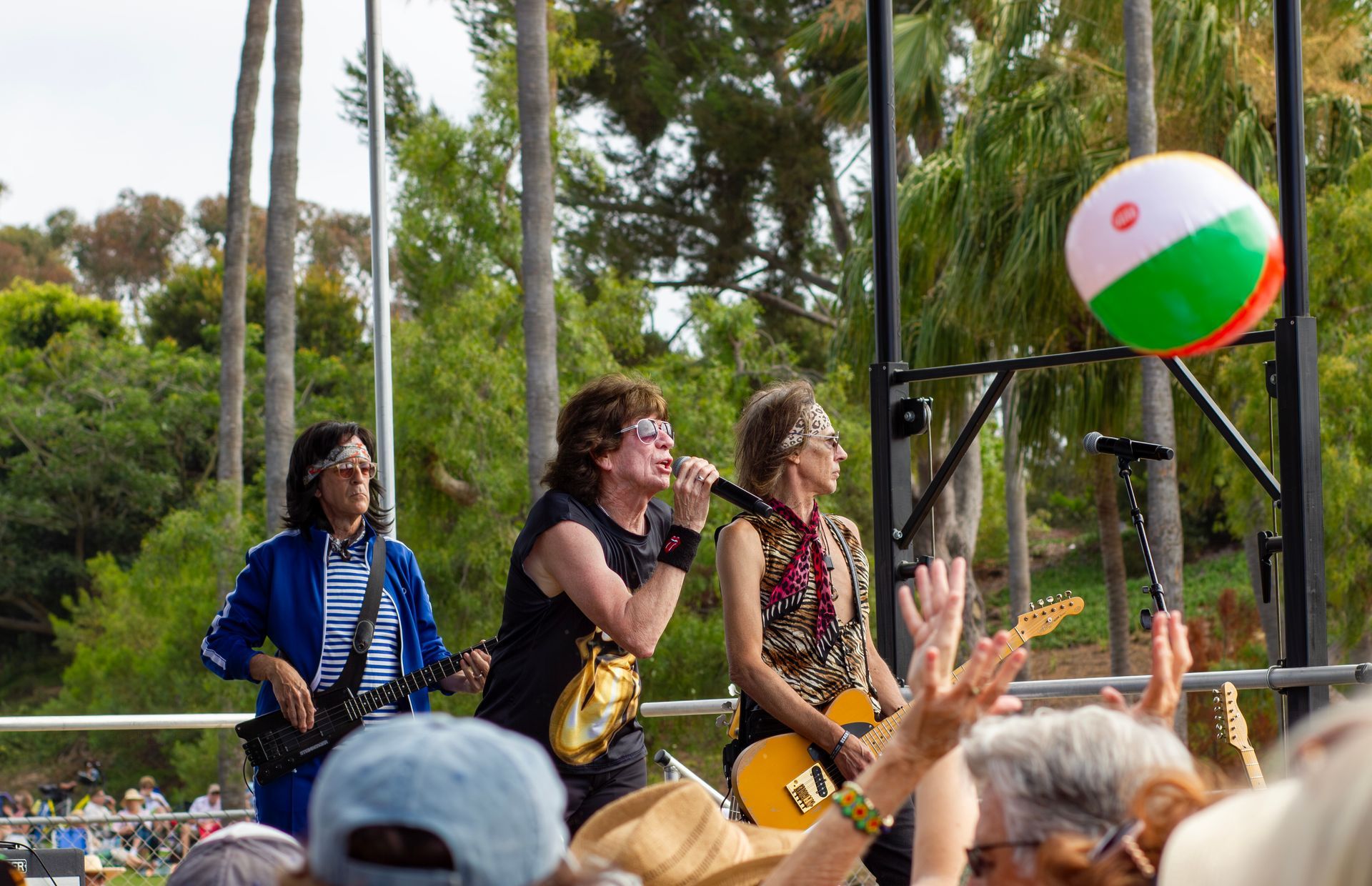 Outdoor band performing on stage with brass instruments and a beach ball floating nearby, crowd in foreground