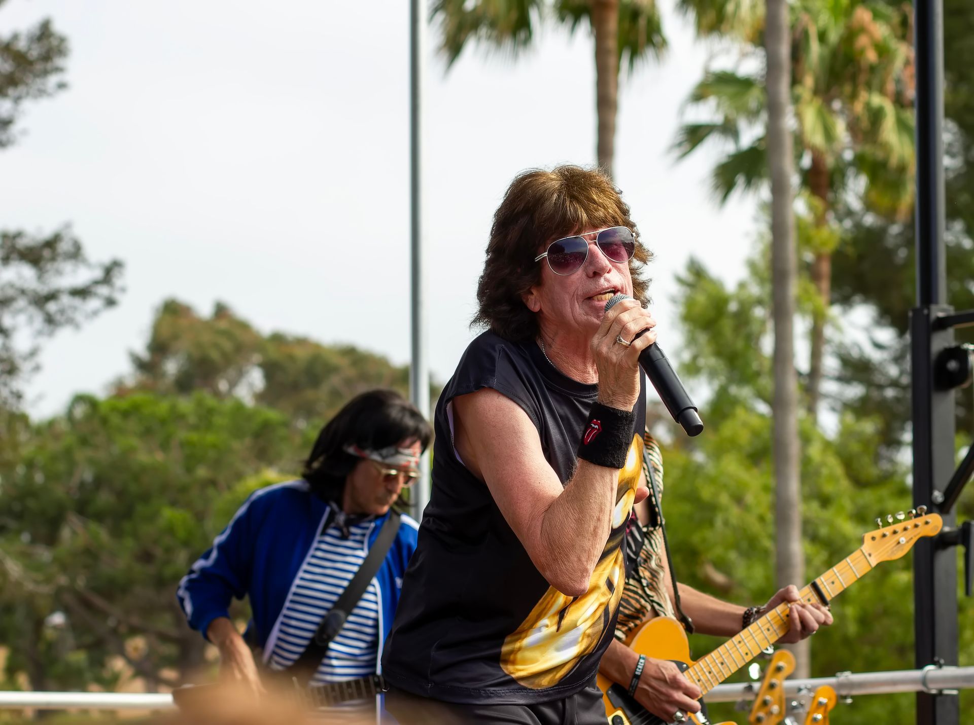 Singer performing outdoors with microphone as guitarist plays behind, on a sunny stage