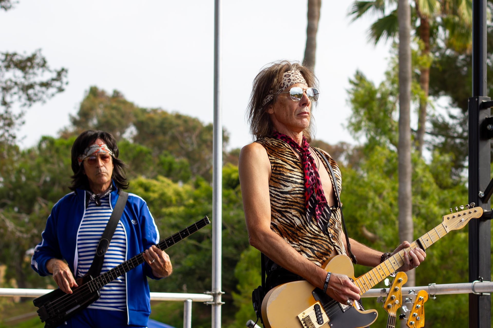 Two musicians playing guitar and bass on an outdoor stage with trees in the background
