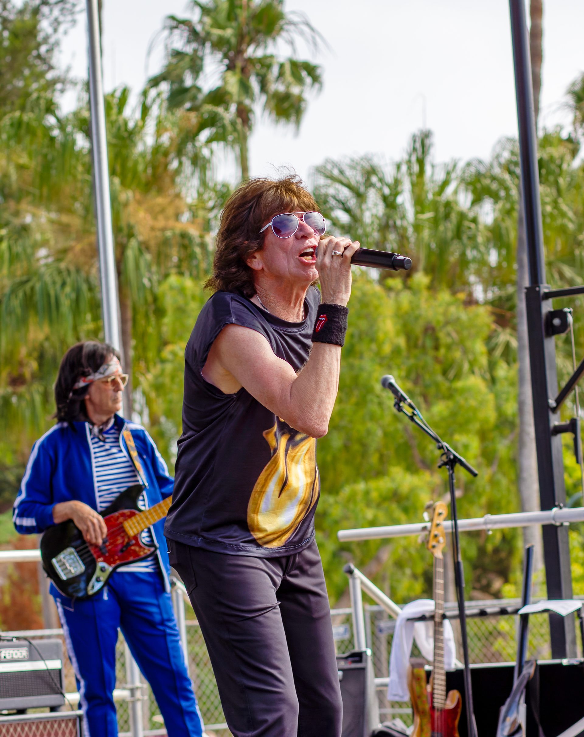 Singer performing on an outdoor stage with guitarist, holding a microphone and wearing sunglasses