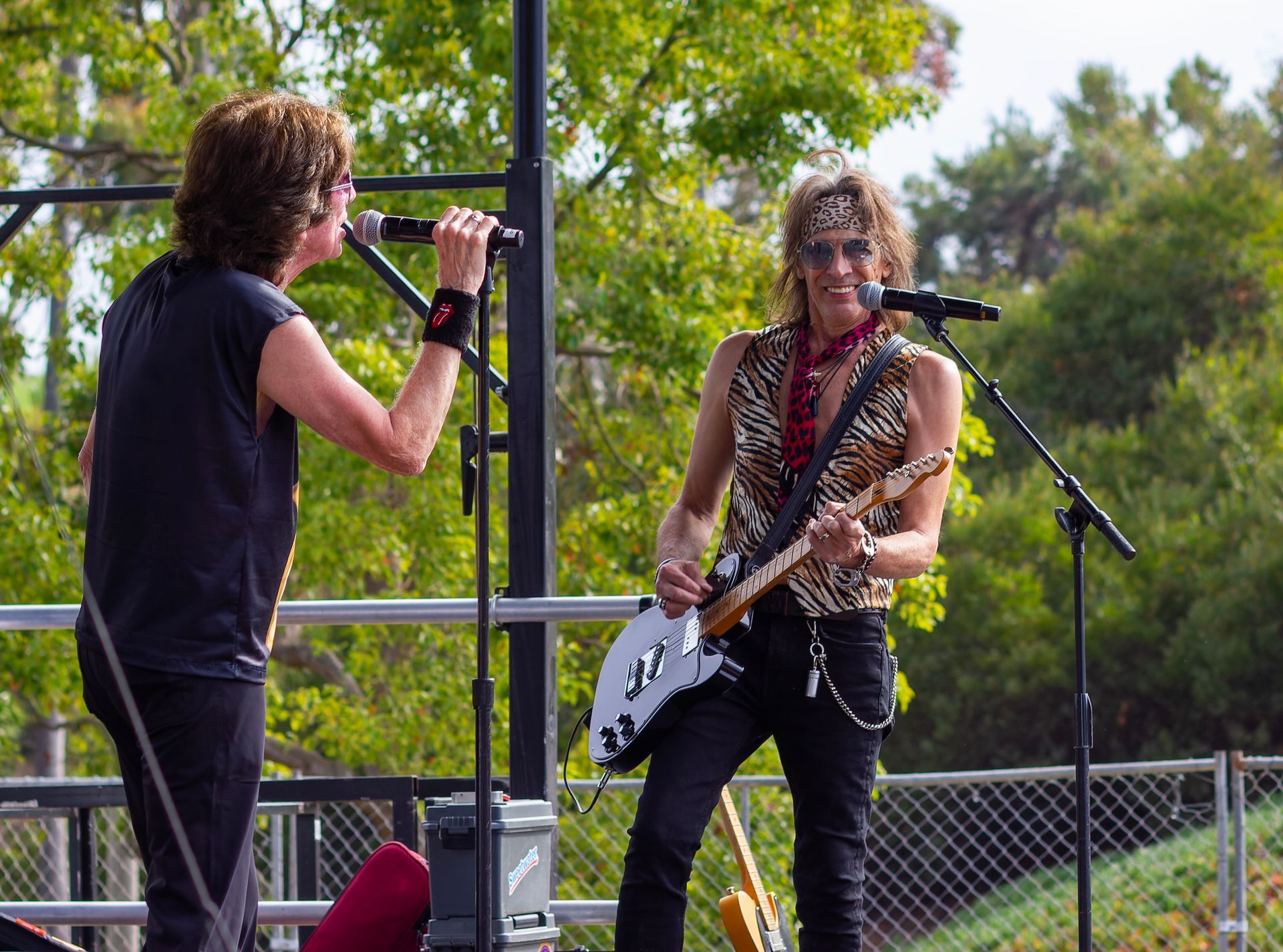 Two musicians perform outdoors, one singing into a mic and one playing electric guitar.
