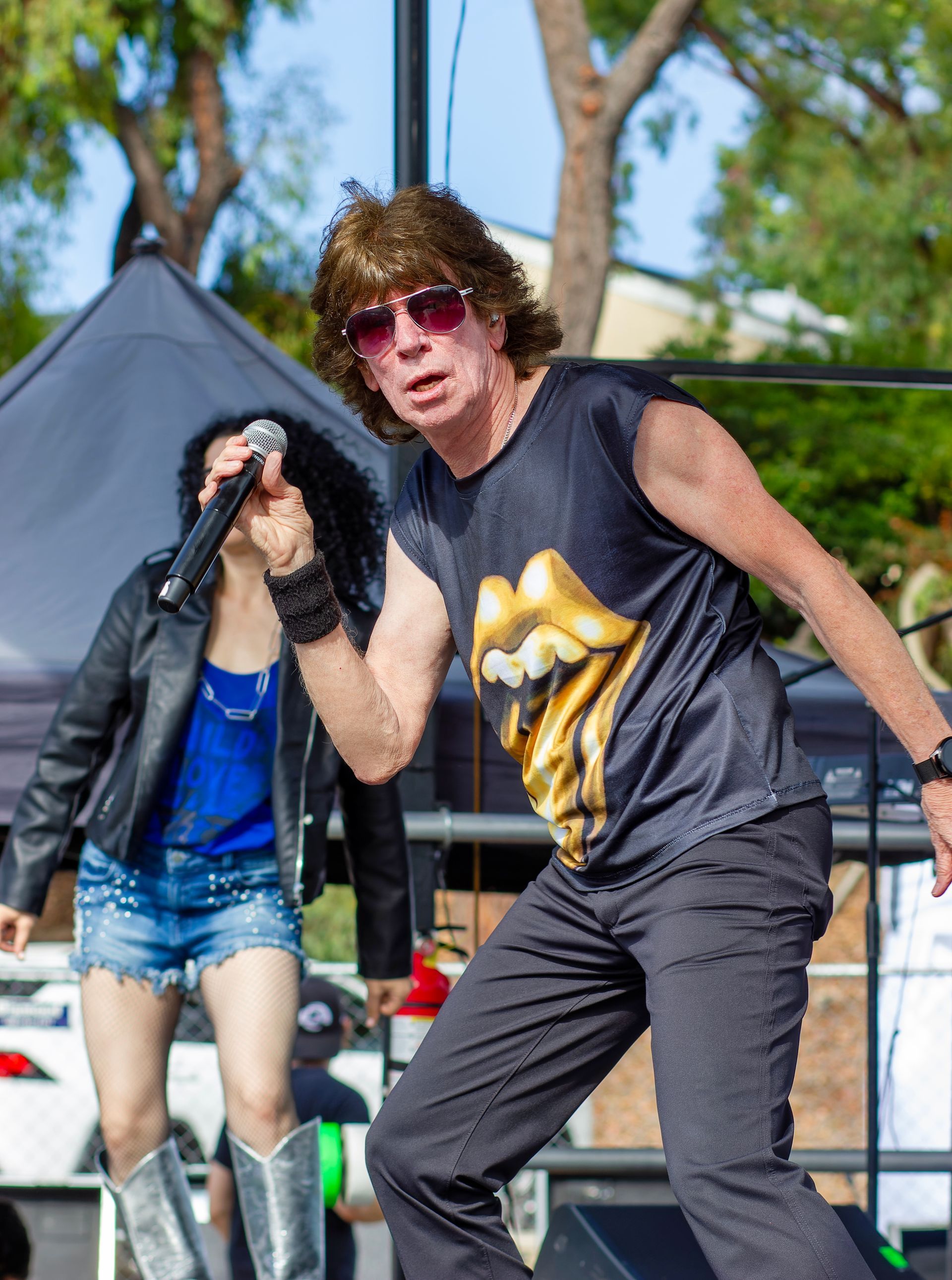 Singer performing outdoors on stage, wearing sunglasses and a sleeveless black shirt, with a guitarist behind.