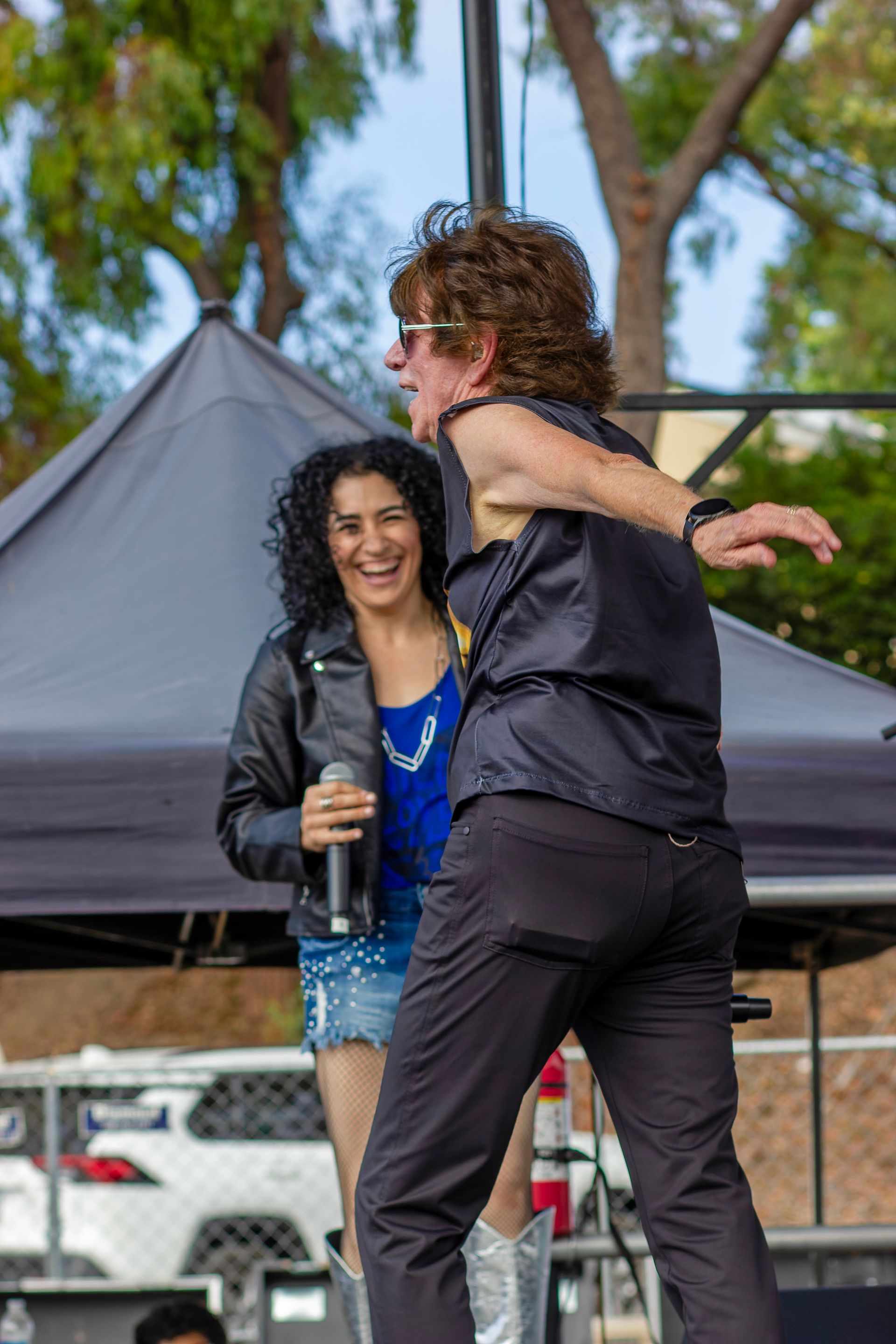 Two performers dancing on an outdoor stage, one holding a microphone, with trees and a tent in the background