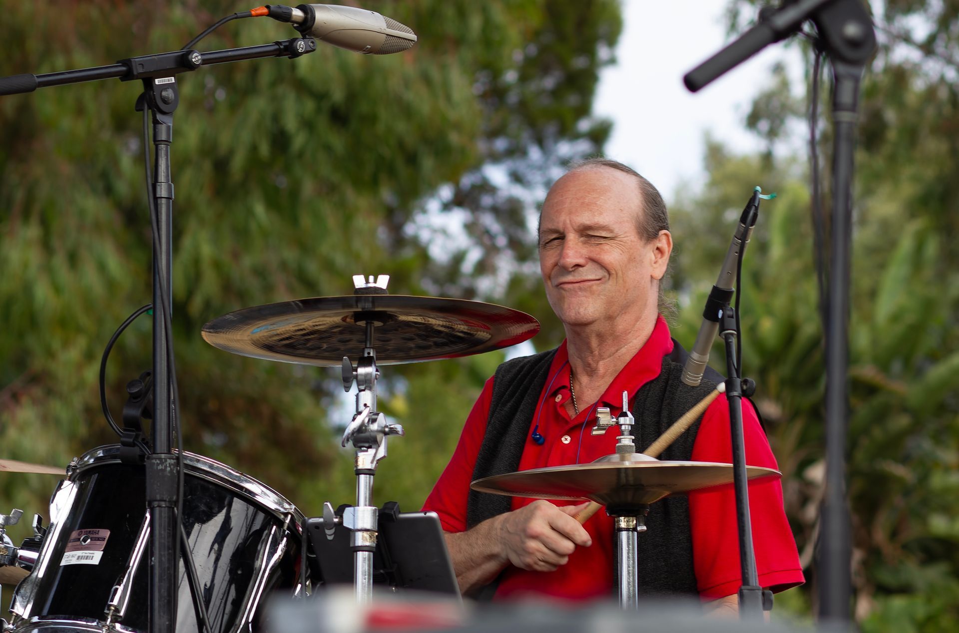Musician in a red shirt playing drums outdoors with microphones overhead