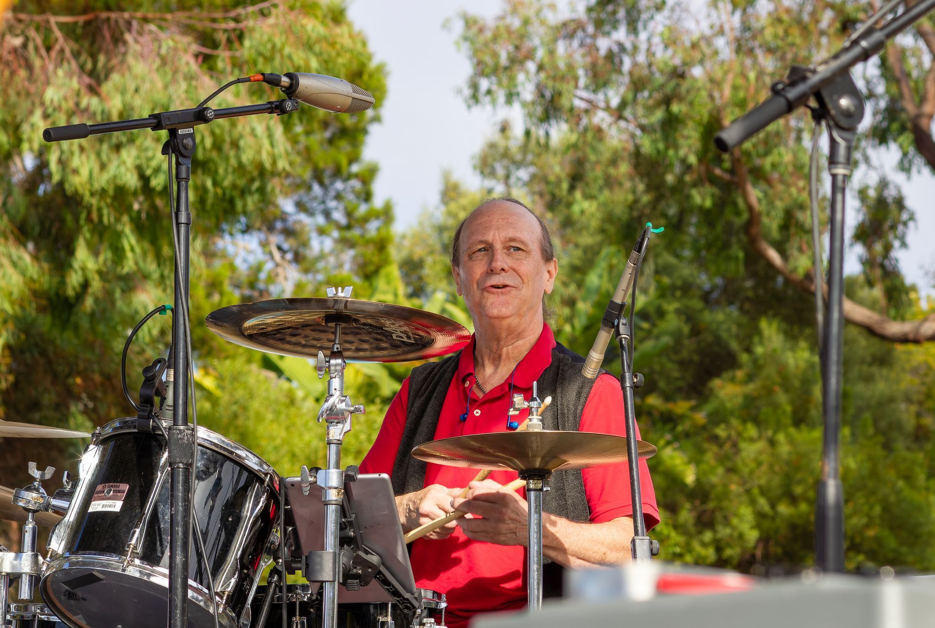 Drummer in a red shirt playing at an outdoor stage with microphones and trees in the background