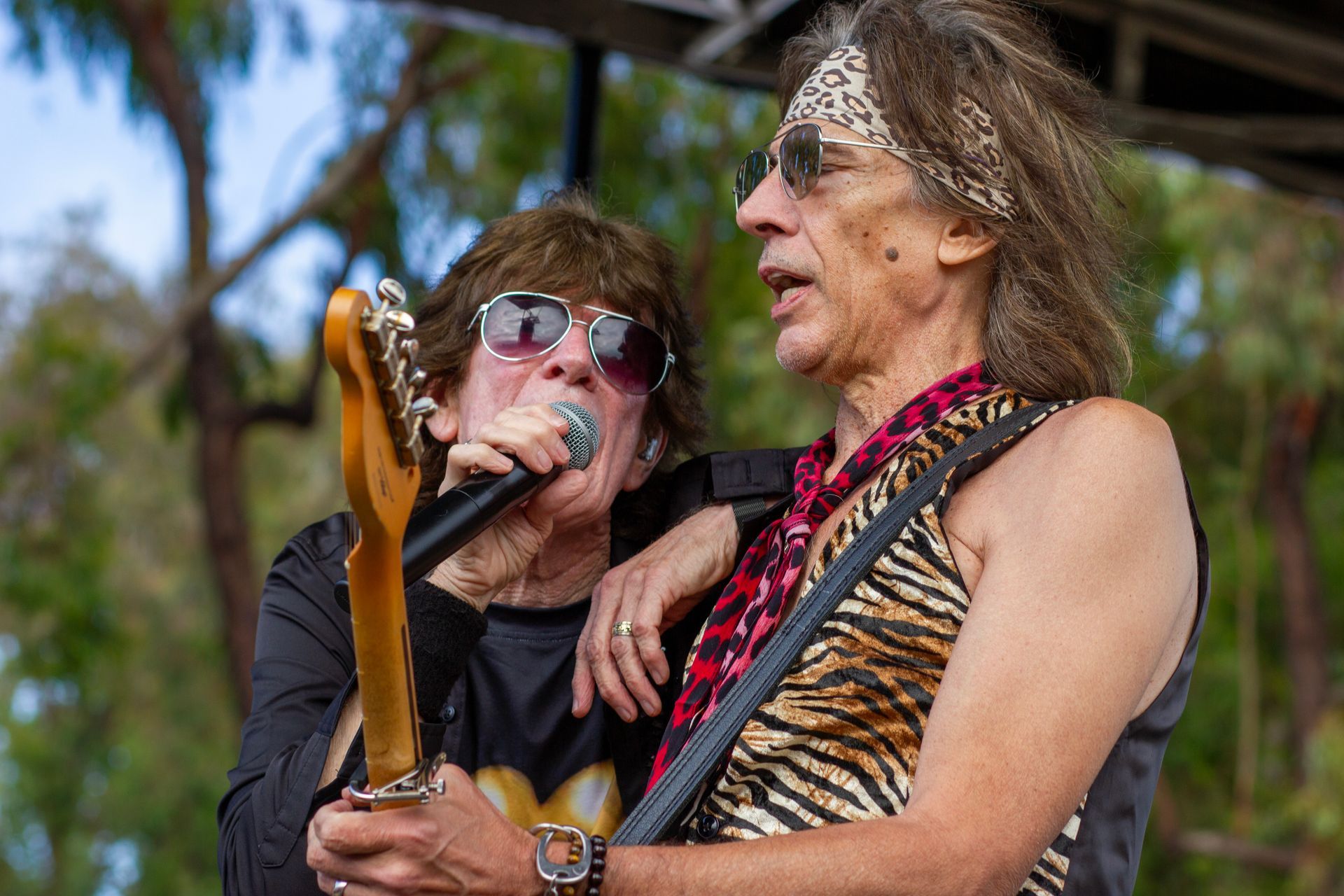 Two musicians singing and playing guitar outdoors, one wearing sunglasses and a zebra-patterned top