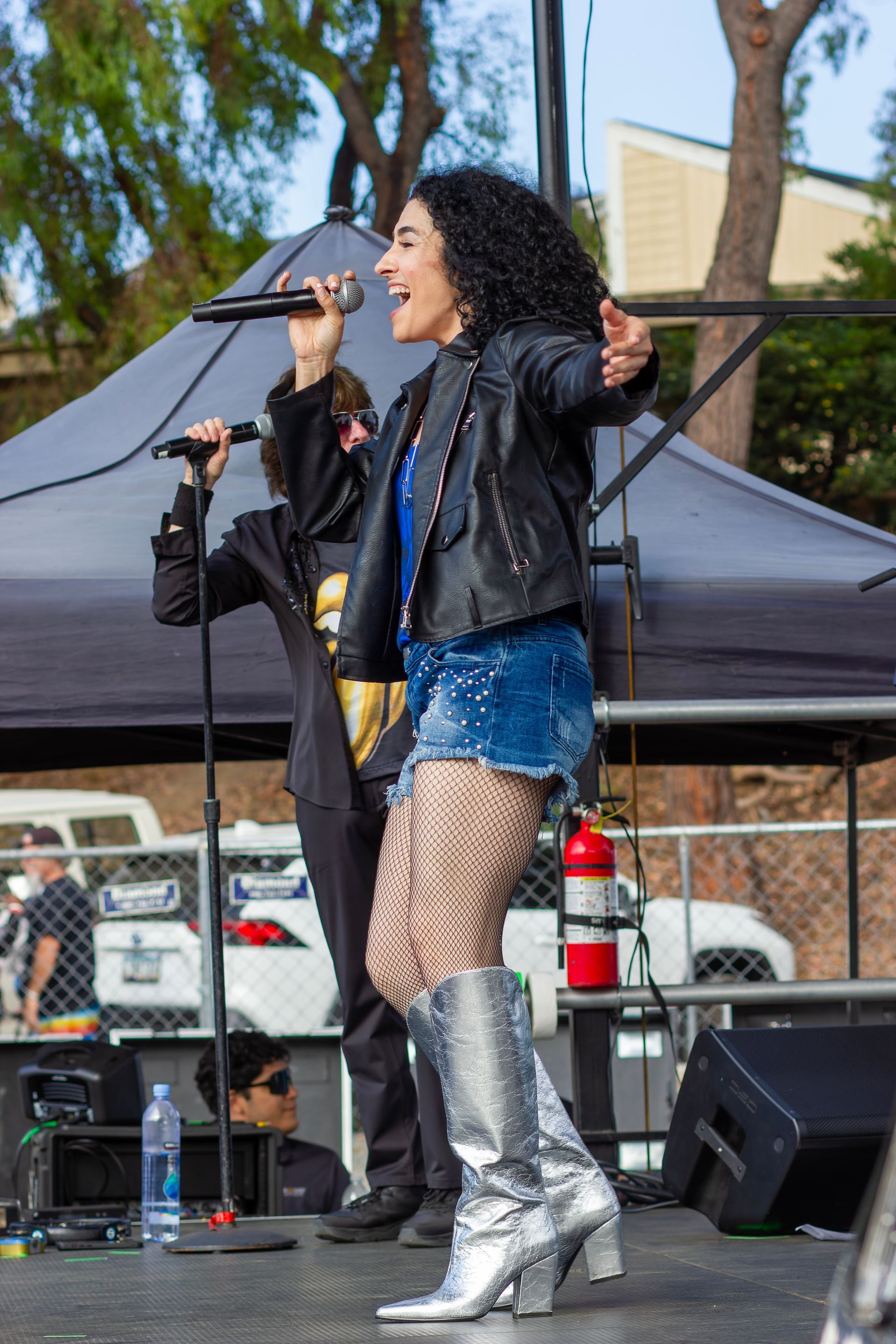 Singer performing on an outdoor stage, wearing a black jacket, blue shorts, fishnet tights, and silver boots.