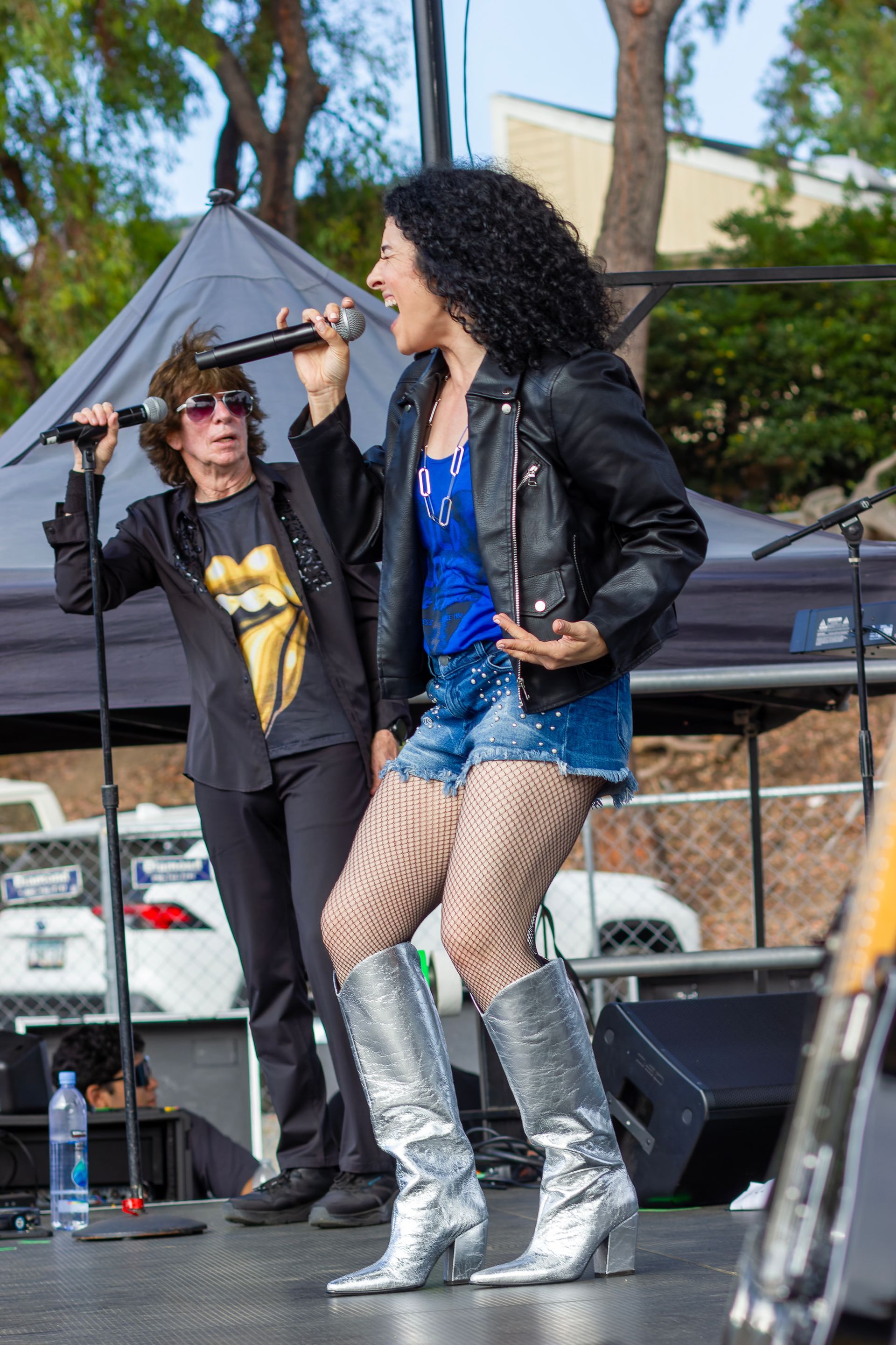 Singer performing on an outdoor stage in a black jacket, blue outfit, and silver boots, with another performer behind.
