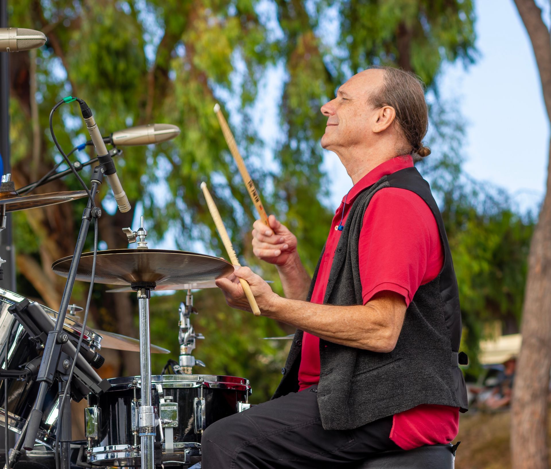 Drummer in a red shirt and black vest playing a drum kit outdoors near trees.