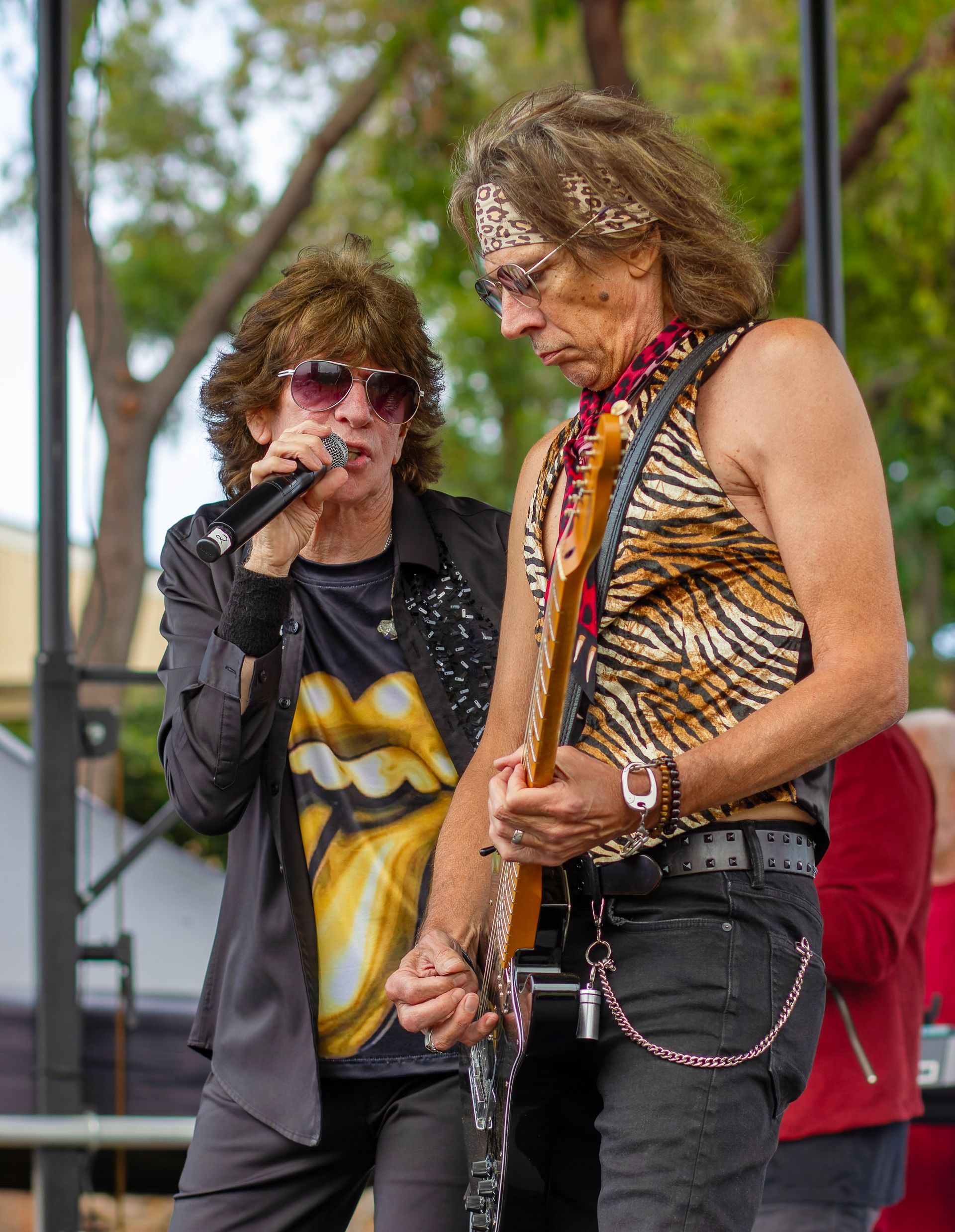 Two musicians performing onstage, one singing and one playing guitar, with an outdoor crowd behind them.