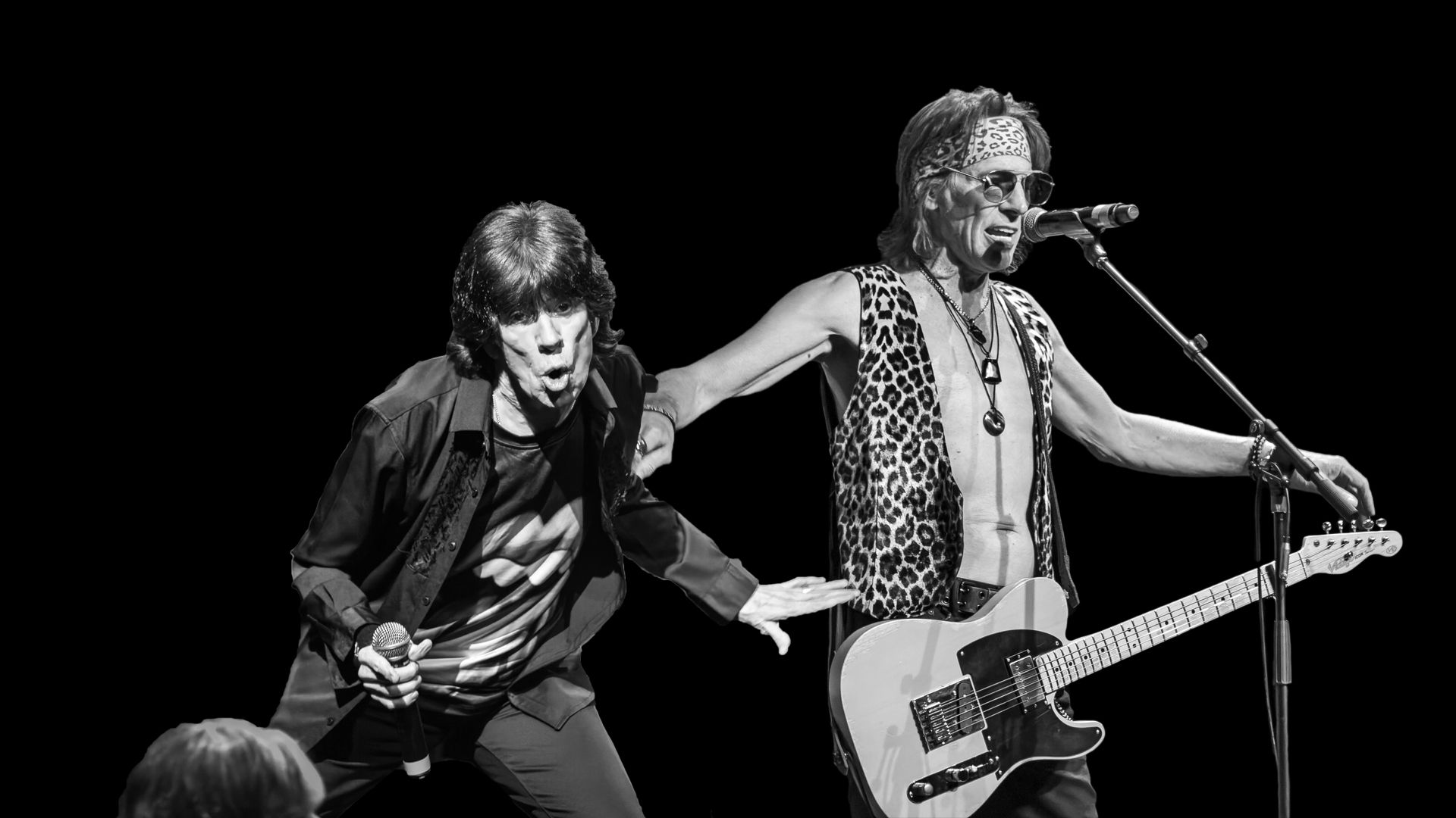 Two musicians onstage, one singing at a microphone and one dancing beside a guitar in black-and-white lighting