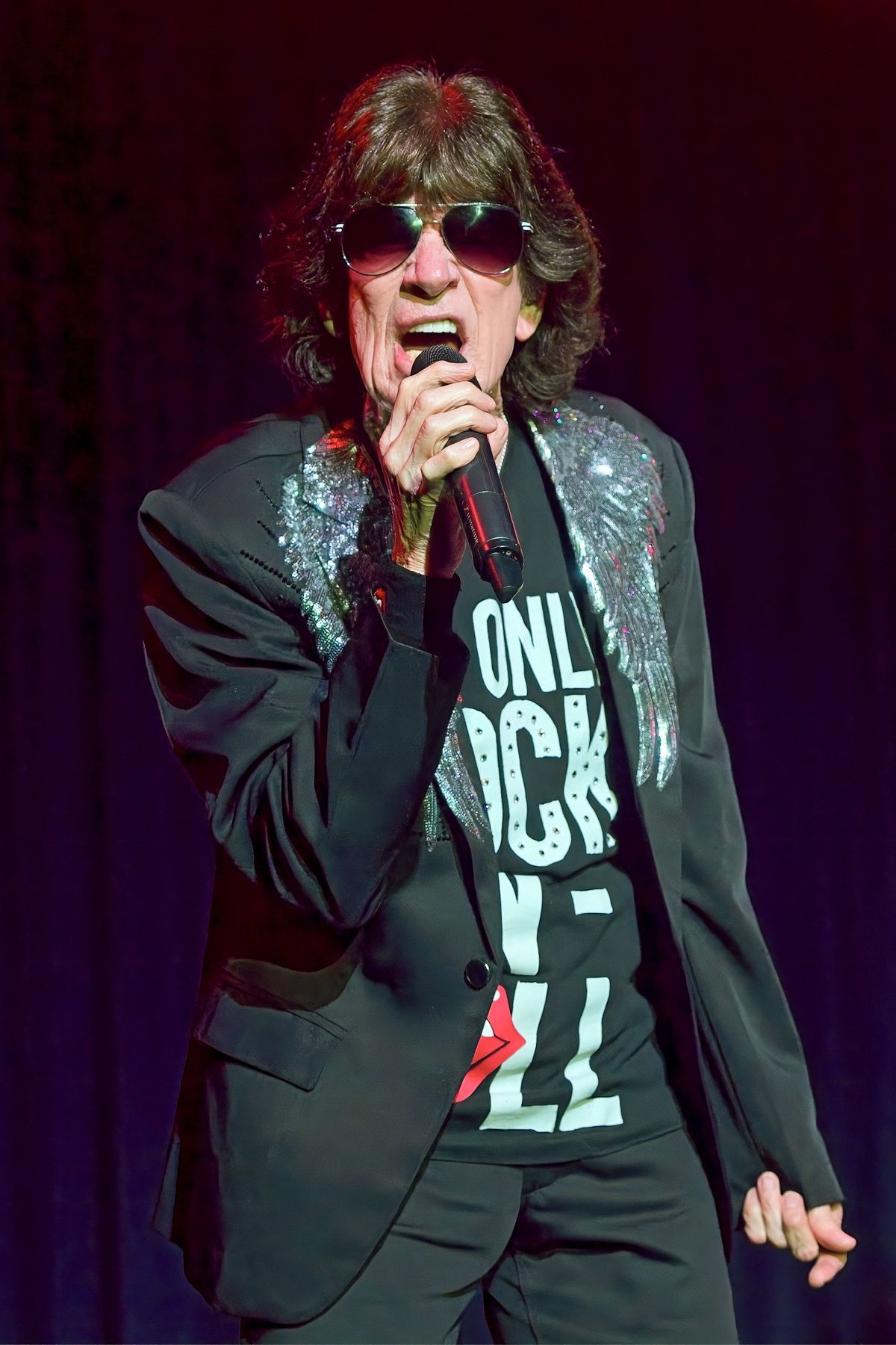 Singer with sunglasses and shaggy hair holding a microphone on a dark stage, wearing a black blazer and graphic T-shirt