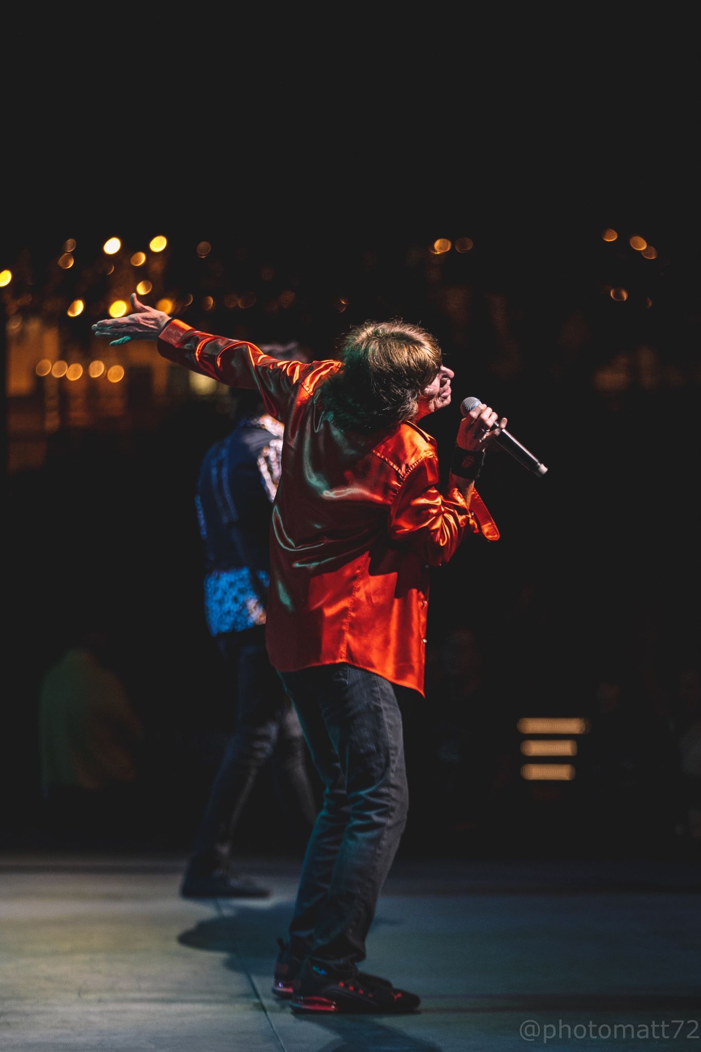 Singer in a red shirt performing onstage with a microphone, under warm stage lights, with a guitarist behind.