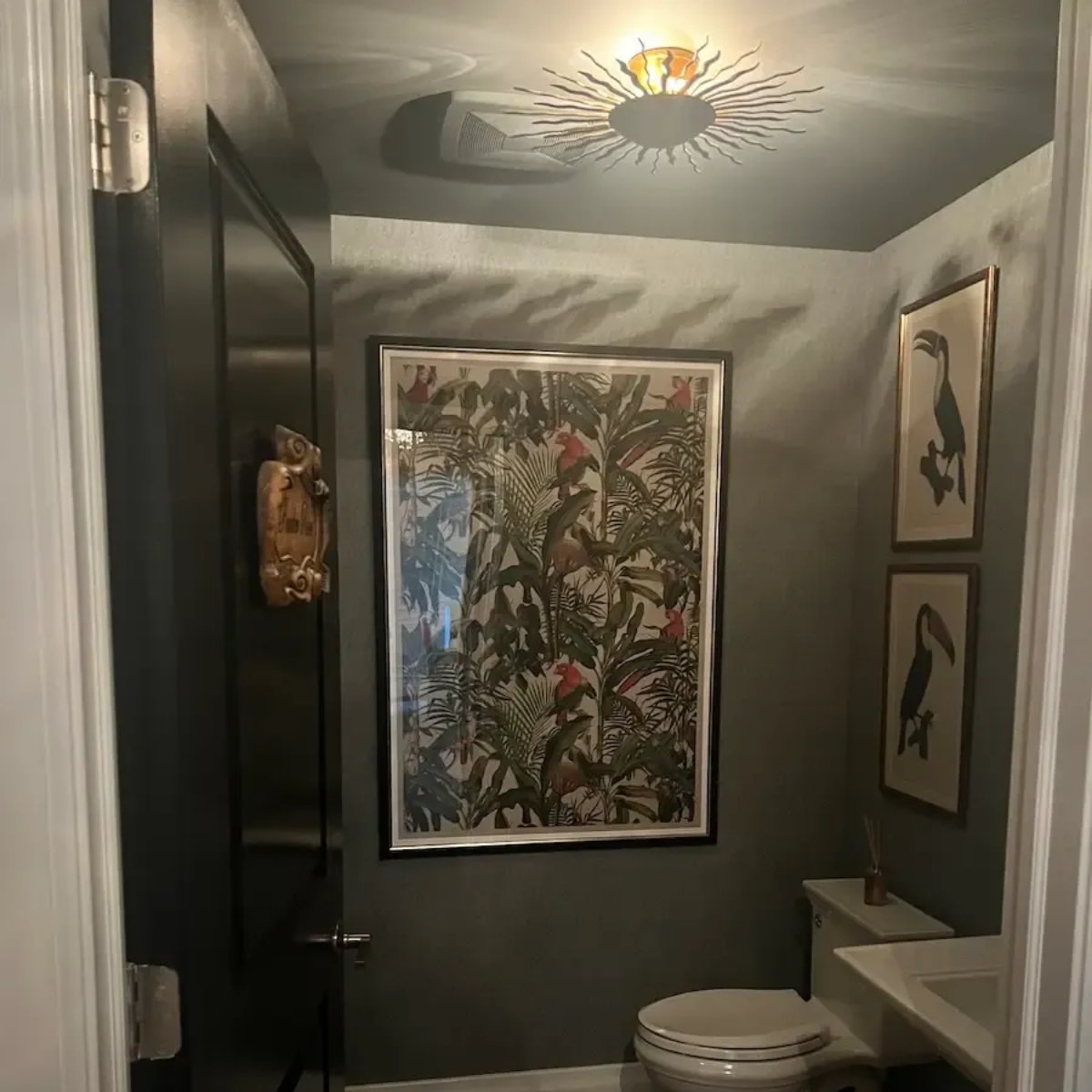 Small, dark-walled bathroom with large art, two bird prints, and gold sunburst ceiling light. Toilet in the center.