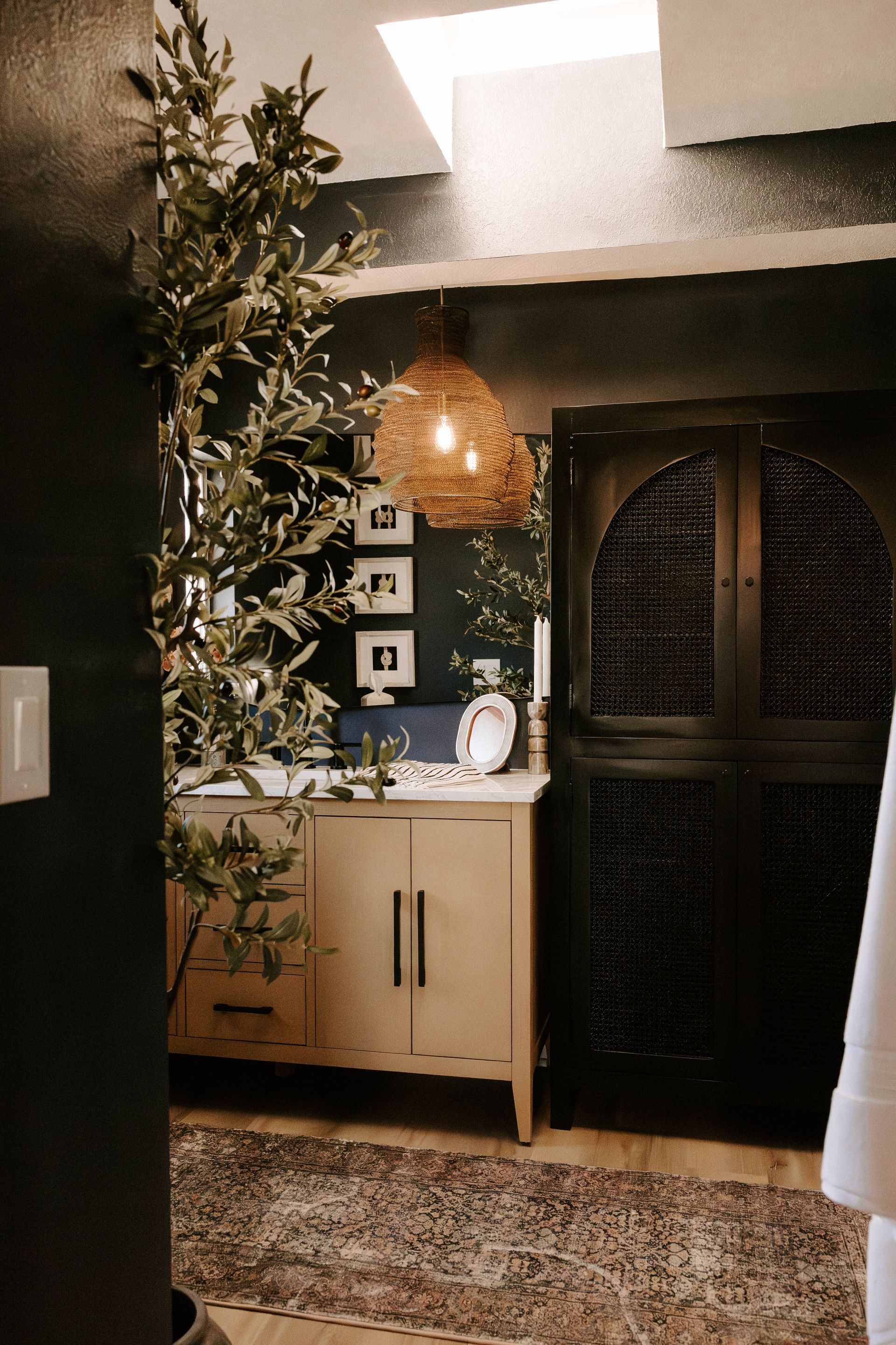 Dark-walled bathroom with light wood vanity, round gold mirrors, and a potted plant.