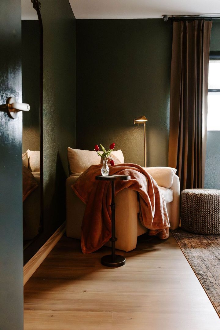Cozy reading nook with armchair, throw blanket, side table, flowers, and floor lamp, set against dark green walls.