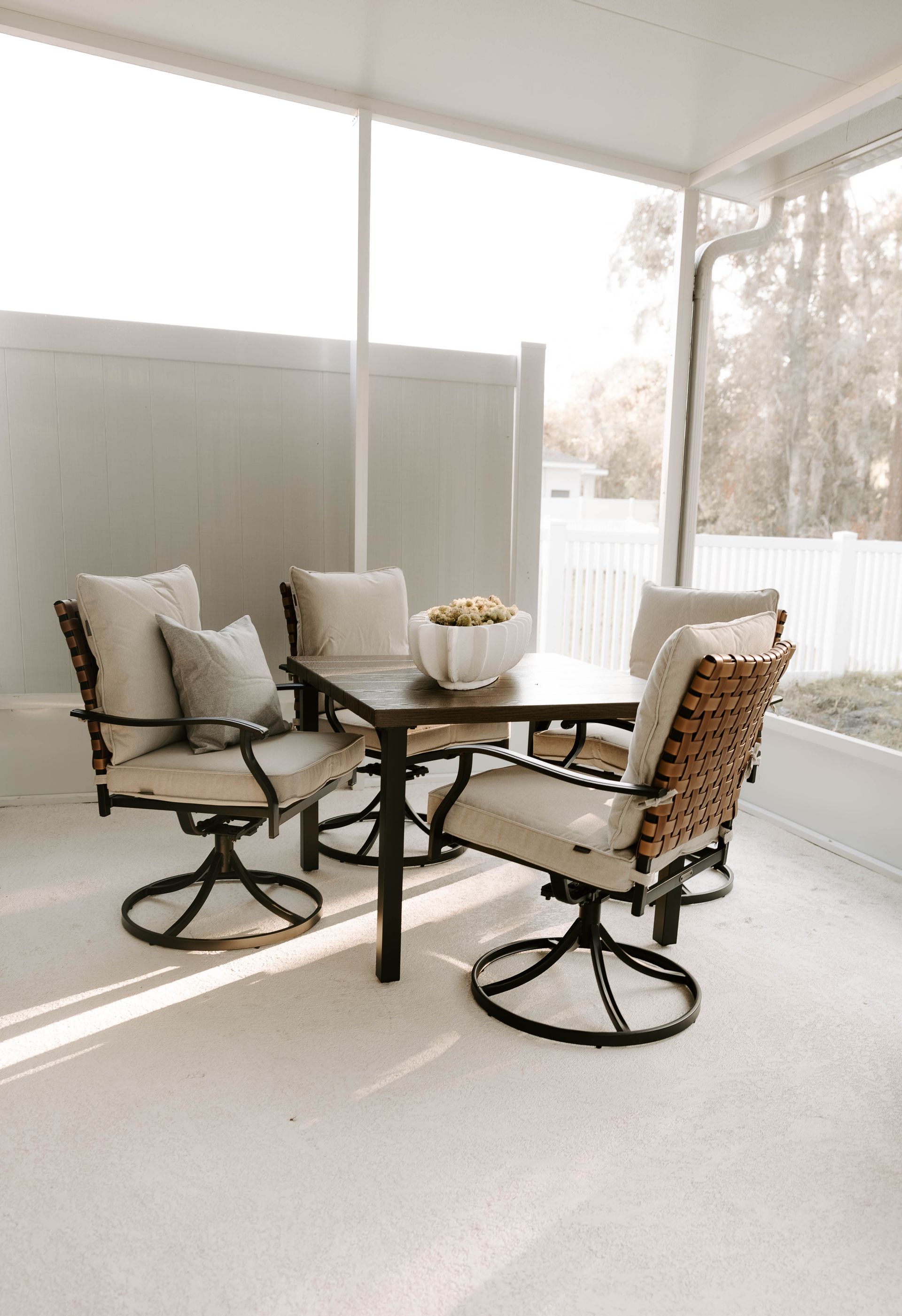 Outdoor dining set in a screened porch: a dark table with four cushioned chairs, white walls, and bright sunlight.