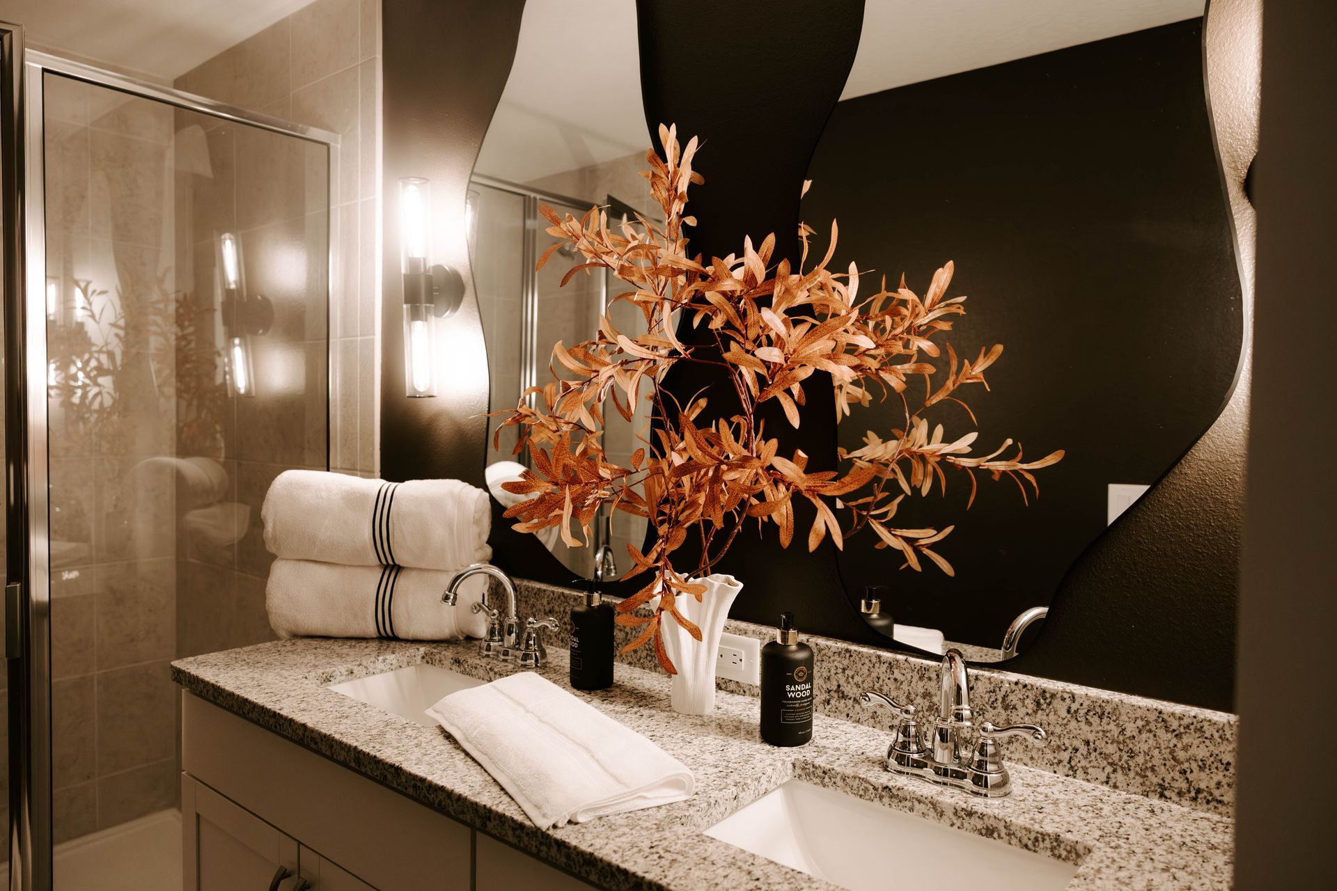 Bathroom with double sinks, granite countertop, two mirrors, decorative branch, and stacked towels.