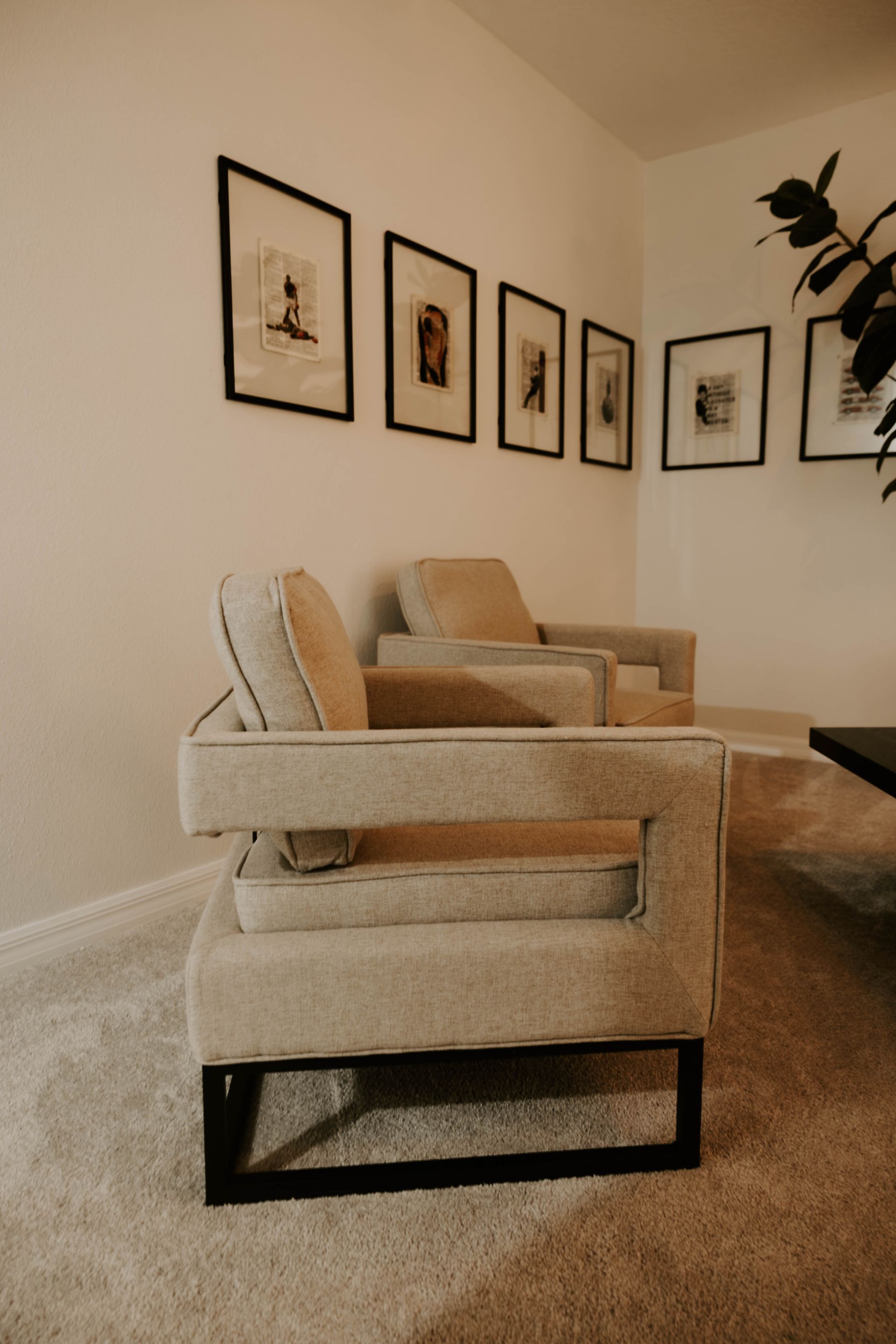Two beige armchairs in a corner with framed artwork on the white wall.