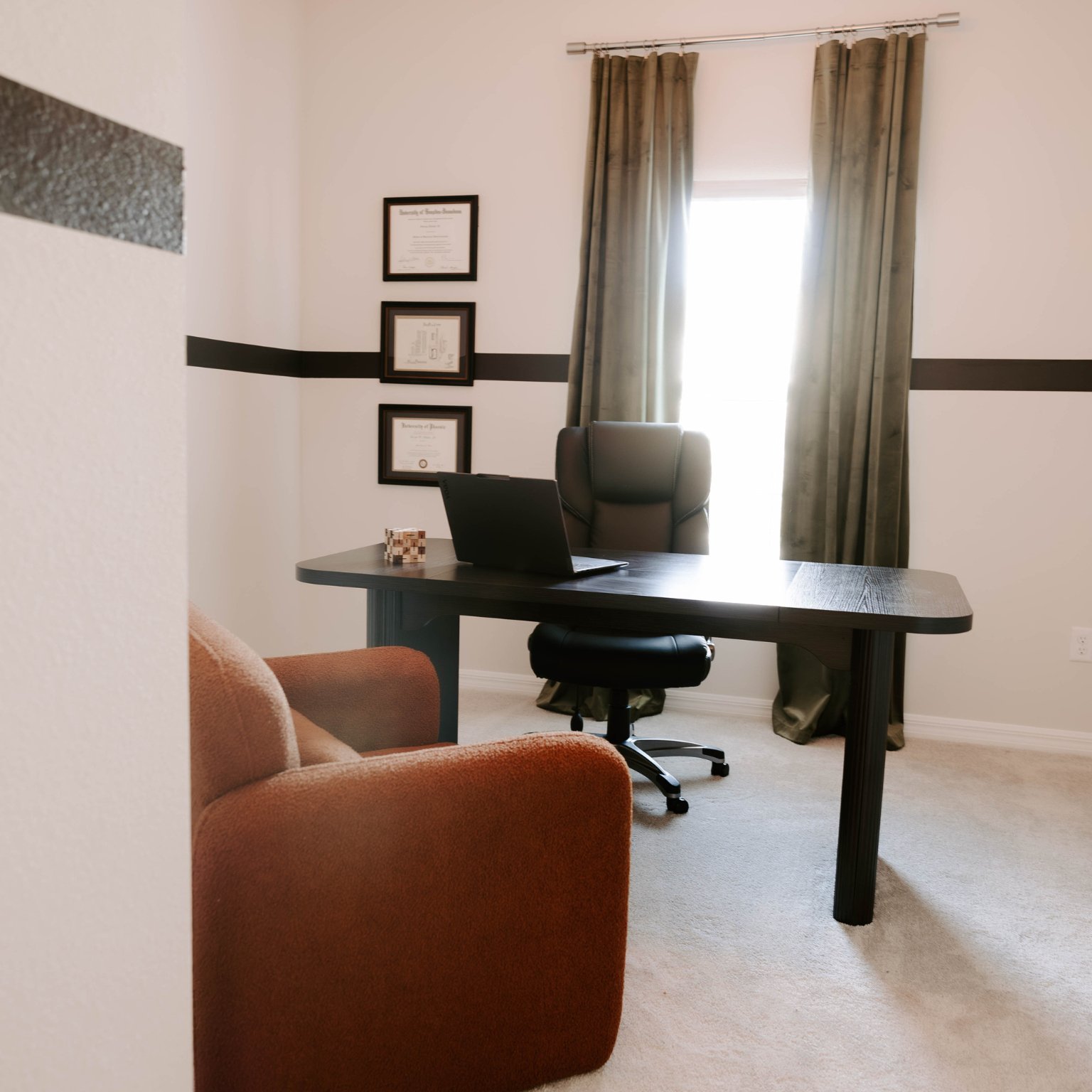 Home office with desk, chair, and diplomas on wall; an armchair is in the foreground.
