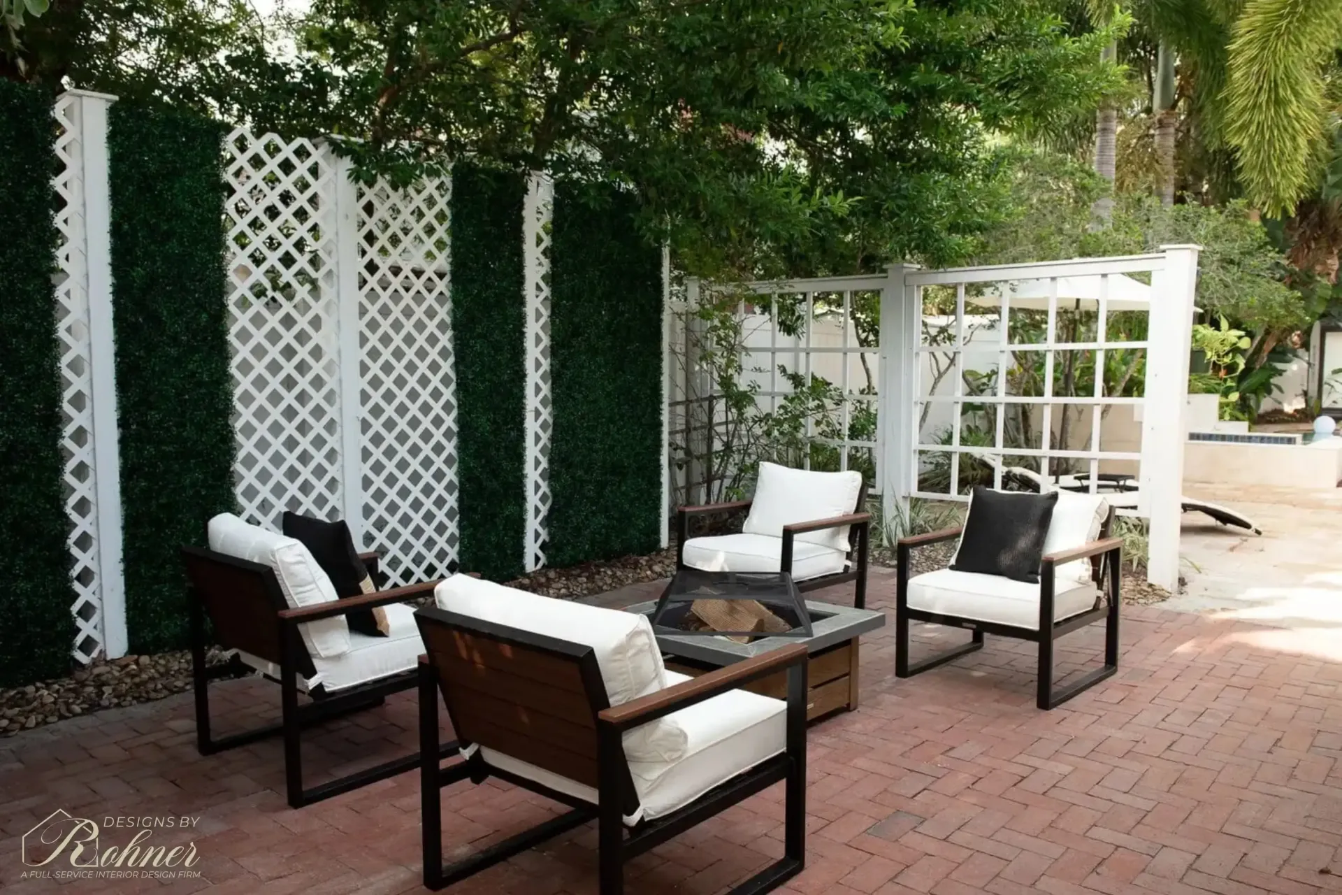 Outdoor patio with seating around a fire pit, green hedge wall, and white trellis.