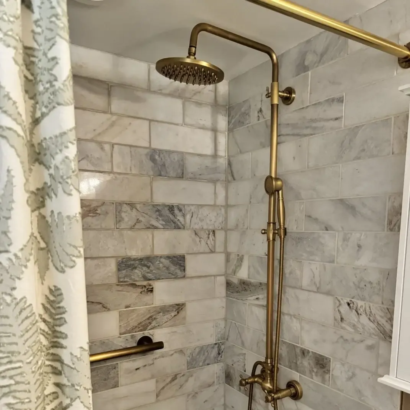 Shower with gold fixtures and marble tile wall, next to a shower curtain with a fern pattern.