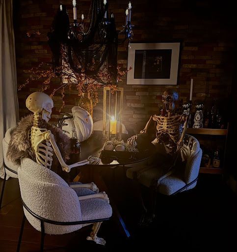 Two skeletons seated at a table, lit by a candle. Halloween-themed decor in a dim setting.