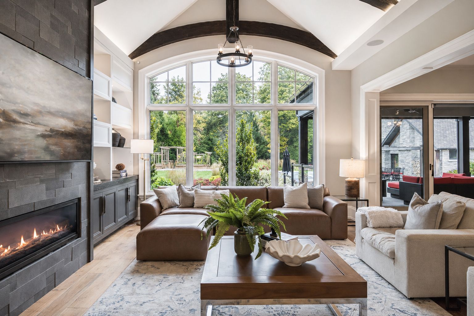 Bright living room with a large arched window, stone fireplace, beige sofas, and wooden beams