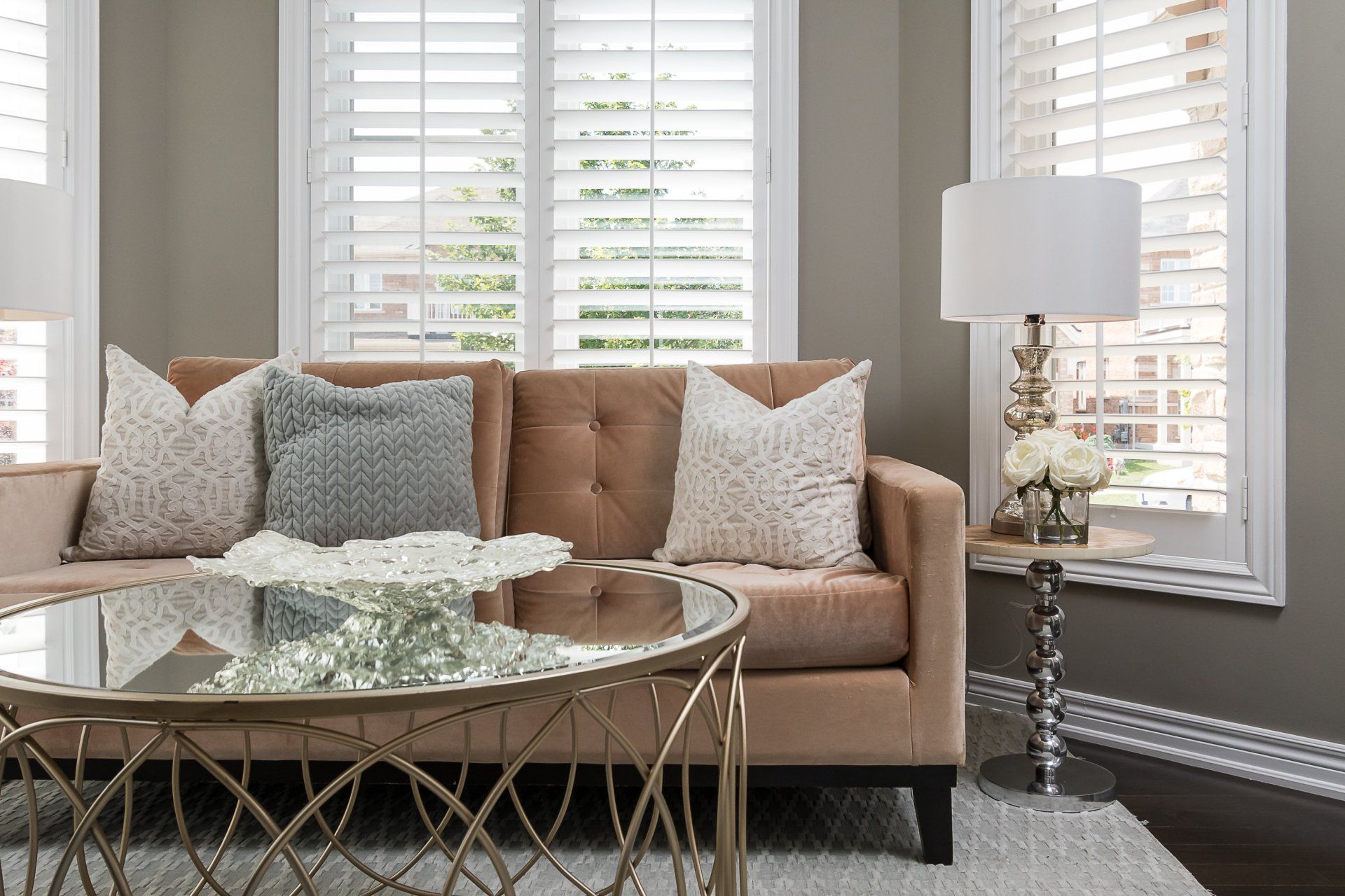 Living room with beige sofa, patterned pillows, glass coffee table, and white shutters