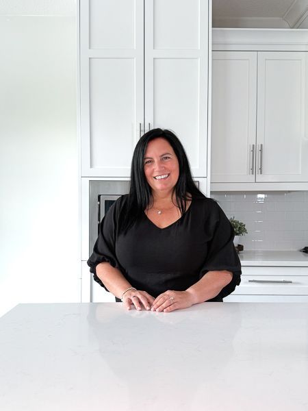 Woman with dark hair smiles in white kitchen, leaning on counter.