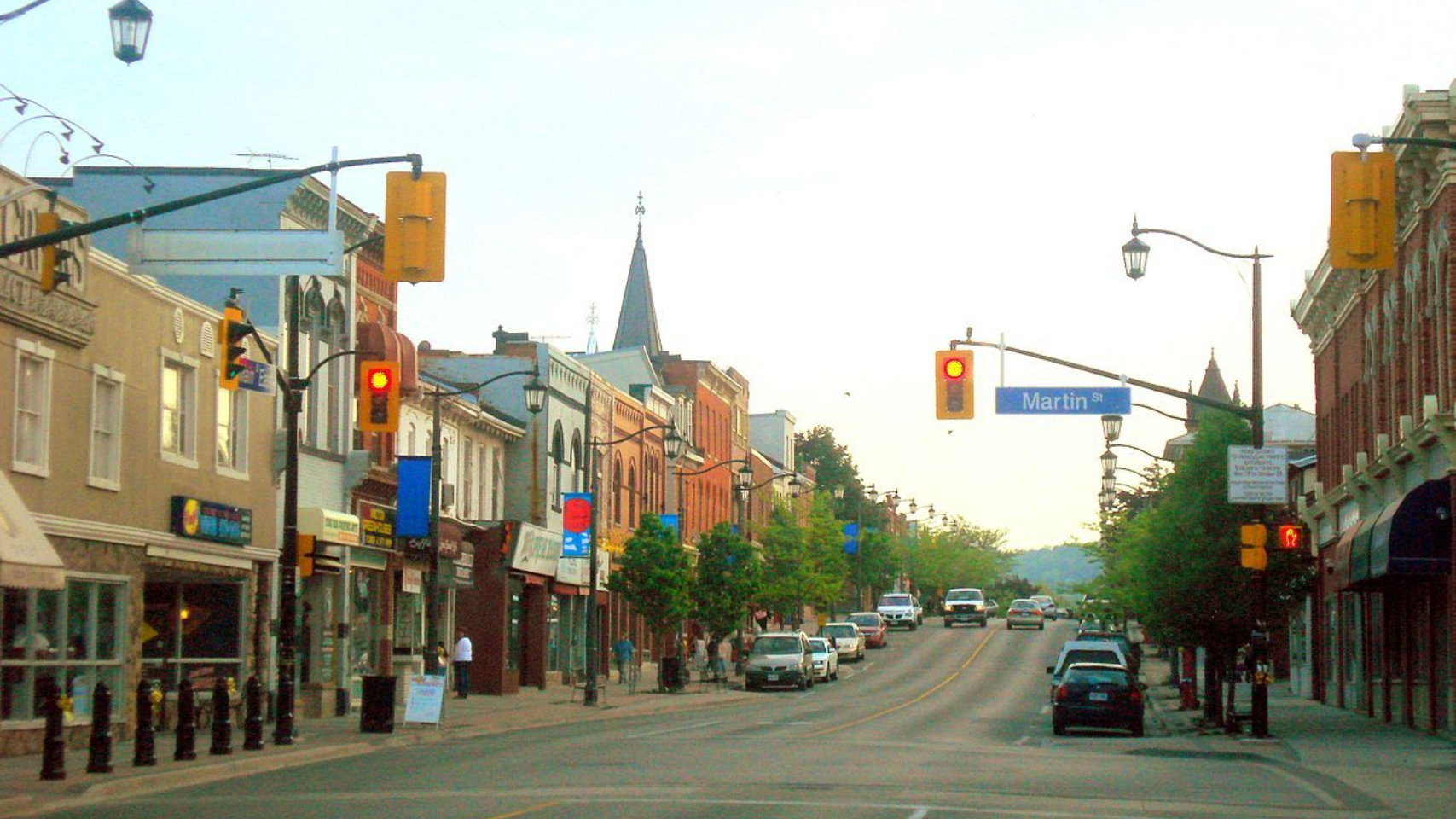 A sunny, quiet main street in a town with brick storefronts, streetlights, traffic signals, and parked cars.