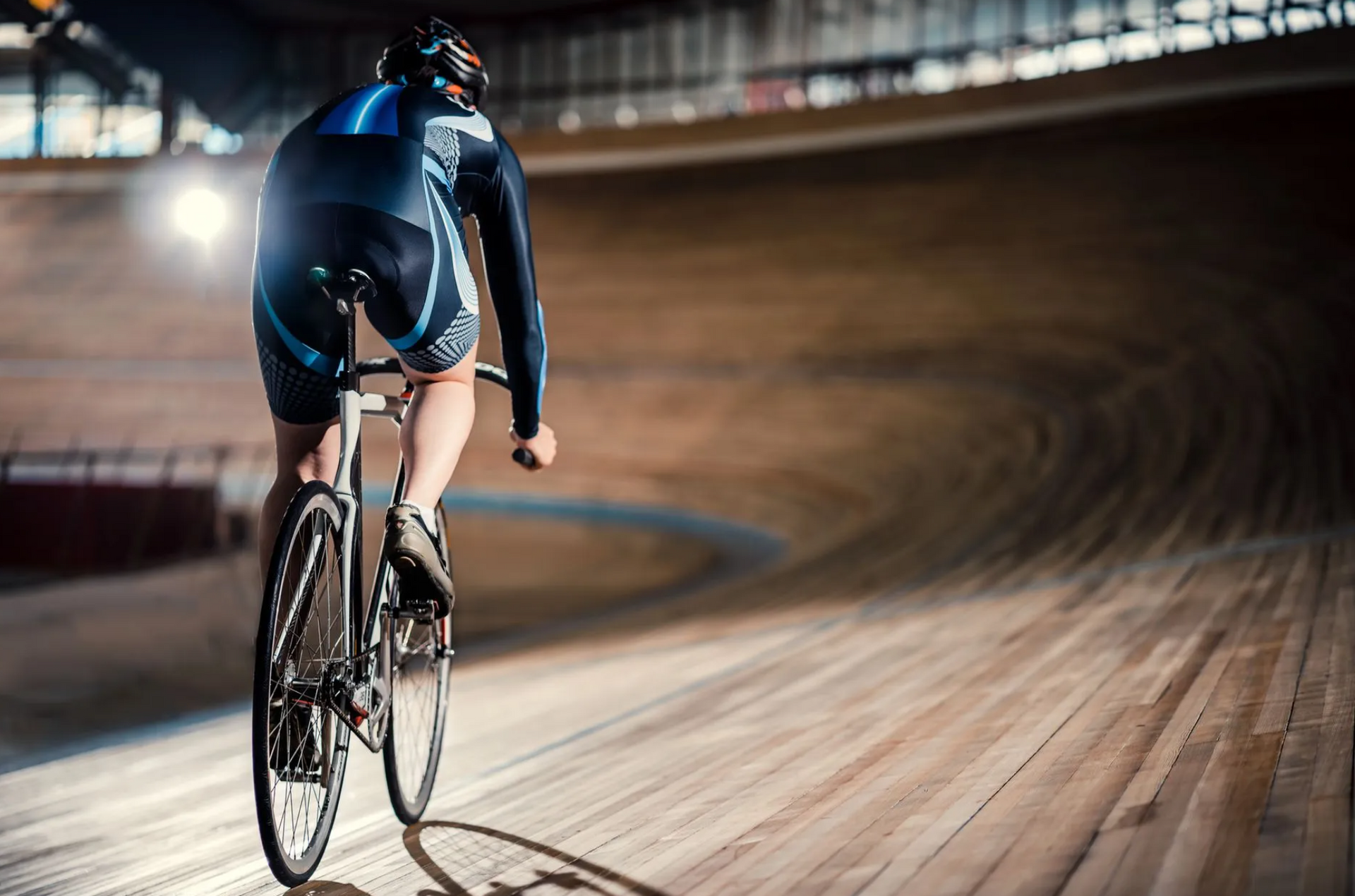 A cyclist rides a track bike on a steep wooden velodrome, seen from behind.