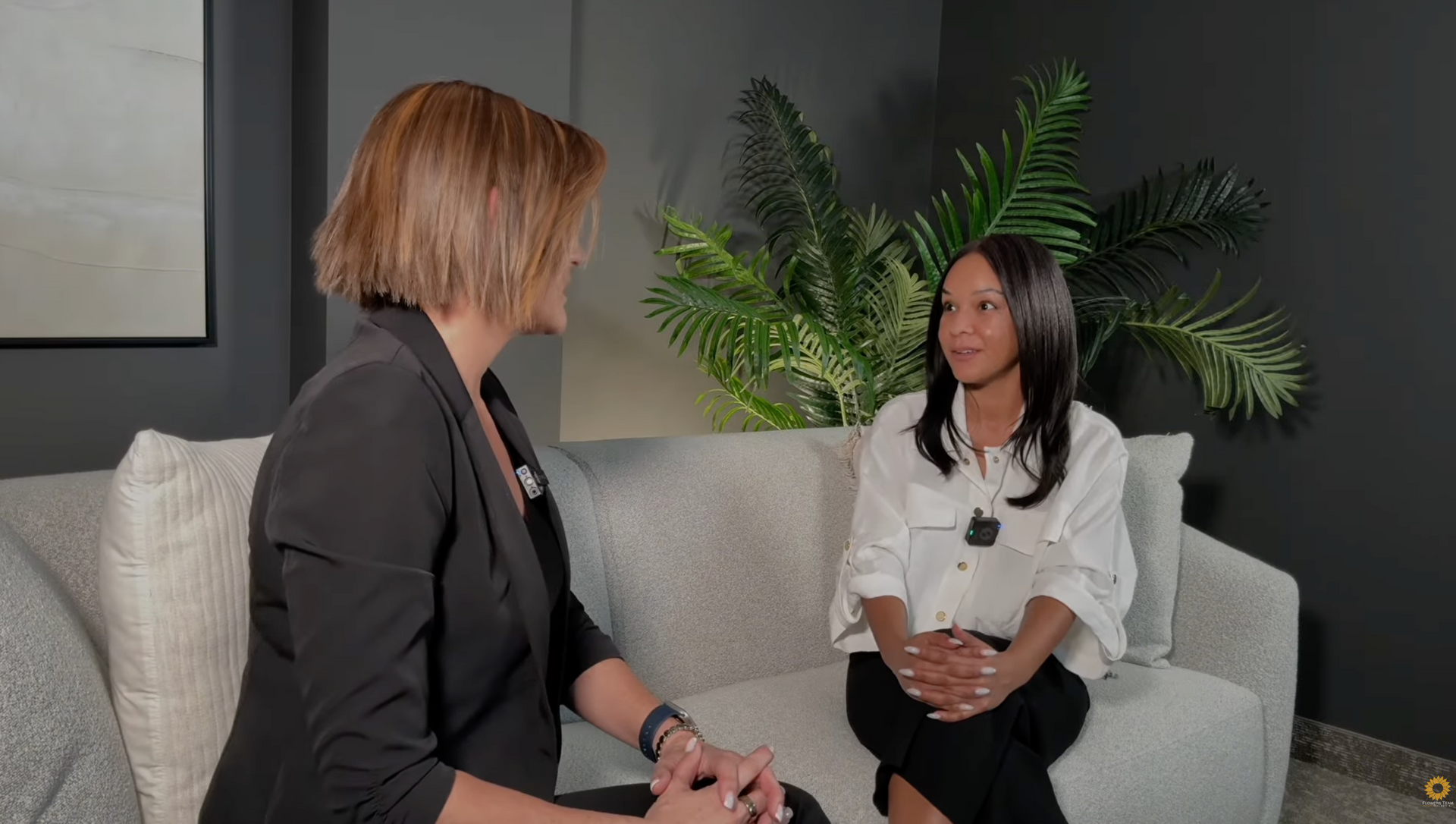 Two women in conversation on a gray sofa in a modern living room with a large plant behind them.