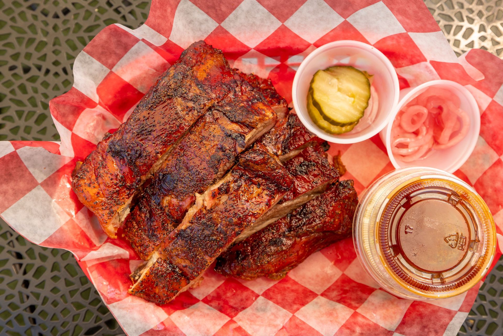 Grilled ribs on checkered paper with pickles, onions, and barbecue sauce cups