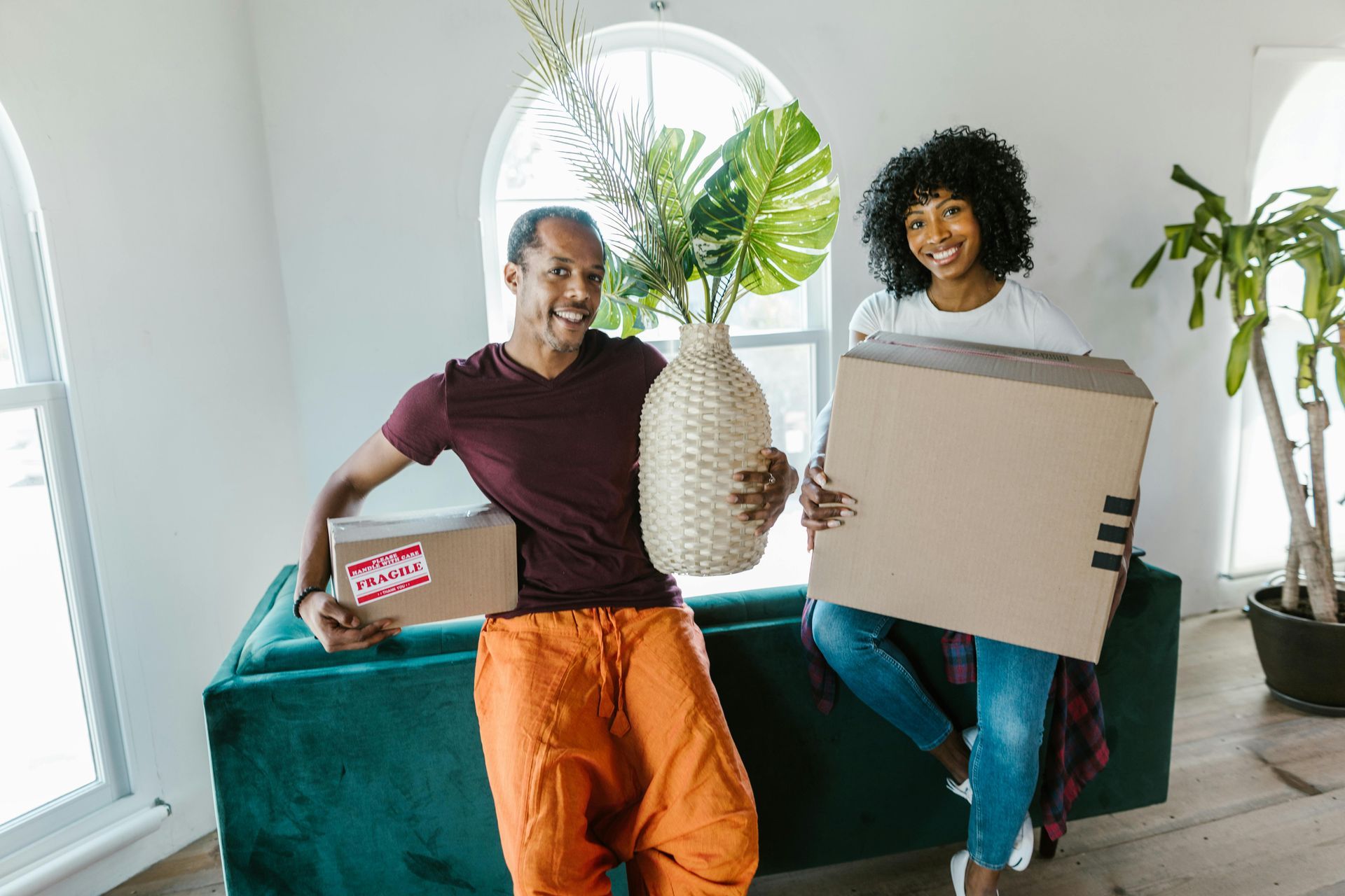 Couple holding boxes and vase, smiling in a room with a green couch and window.