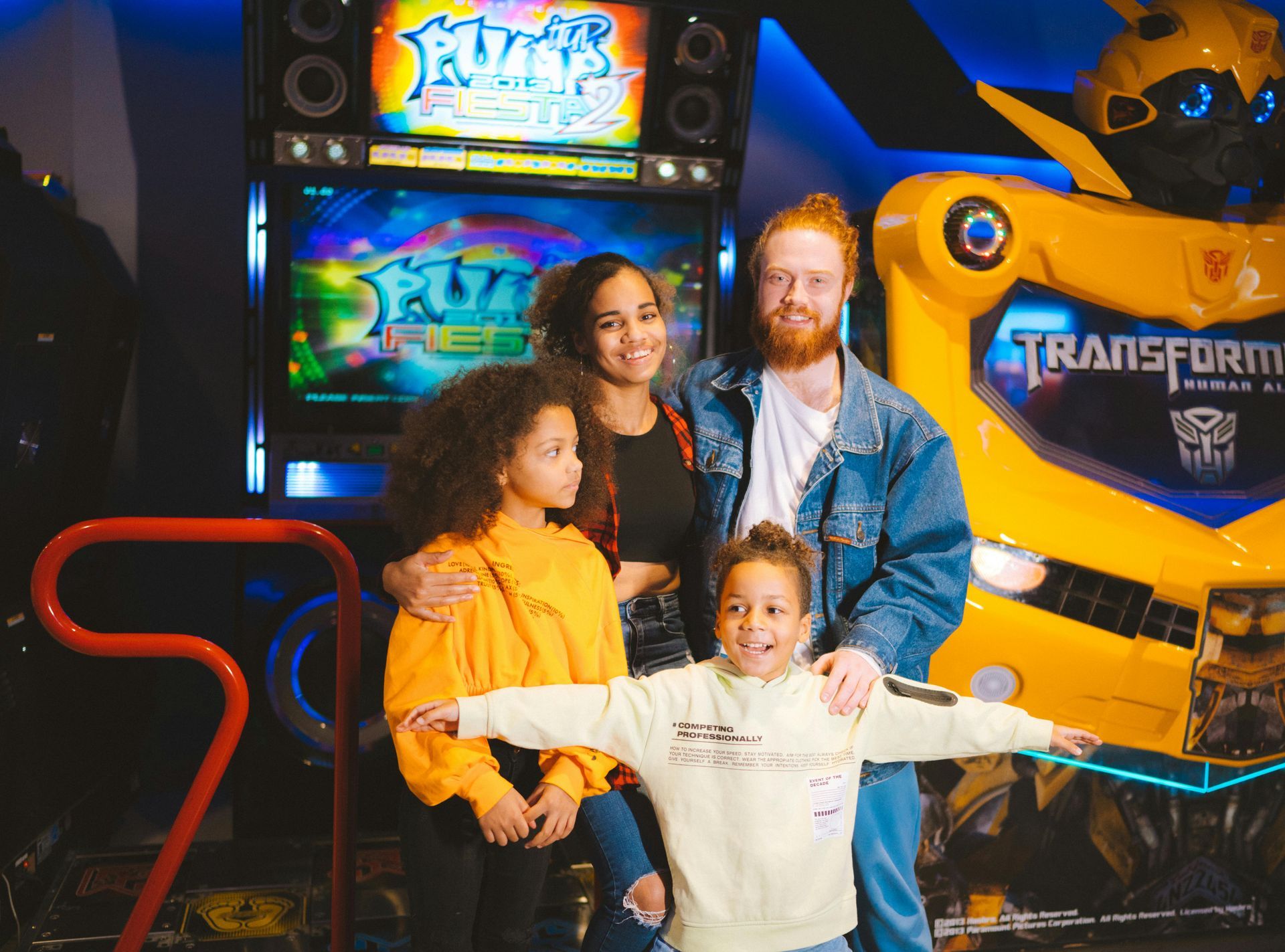 Family poses for photo in front of arcade games, including a yellow Transformer robot.