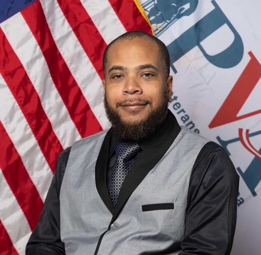 A man with a beard is sitting in front of an american flag