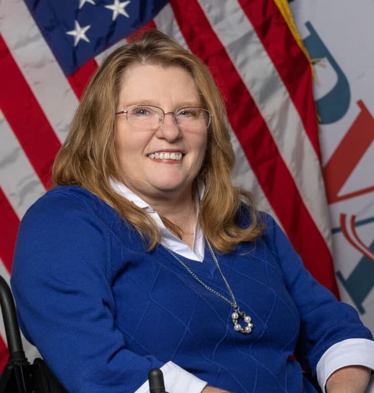 A woman in a wheelchair is smiling in front of an american flag