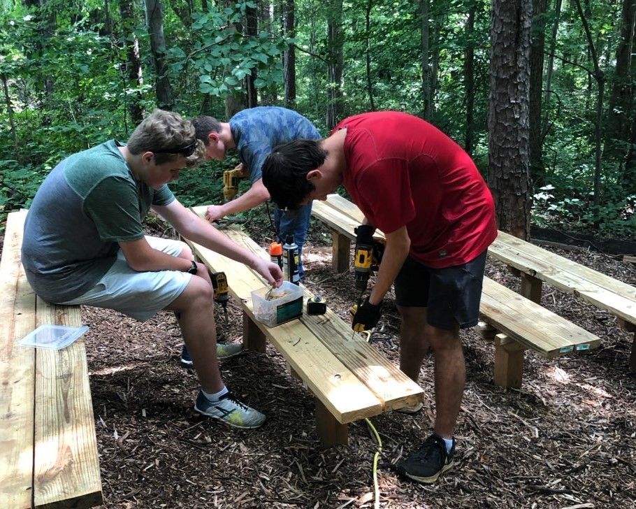 Three men are working on a wooden picnic table in the woods.