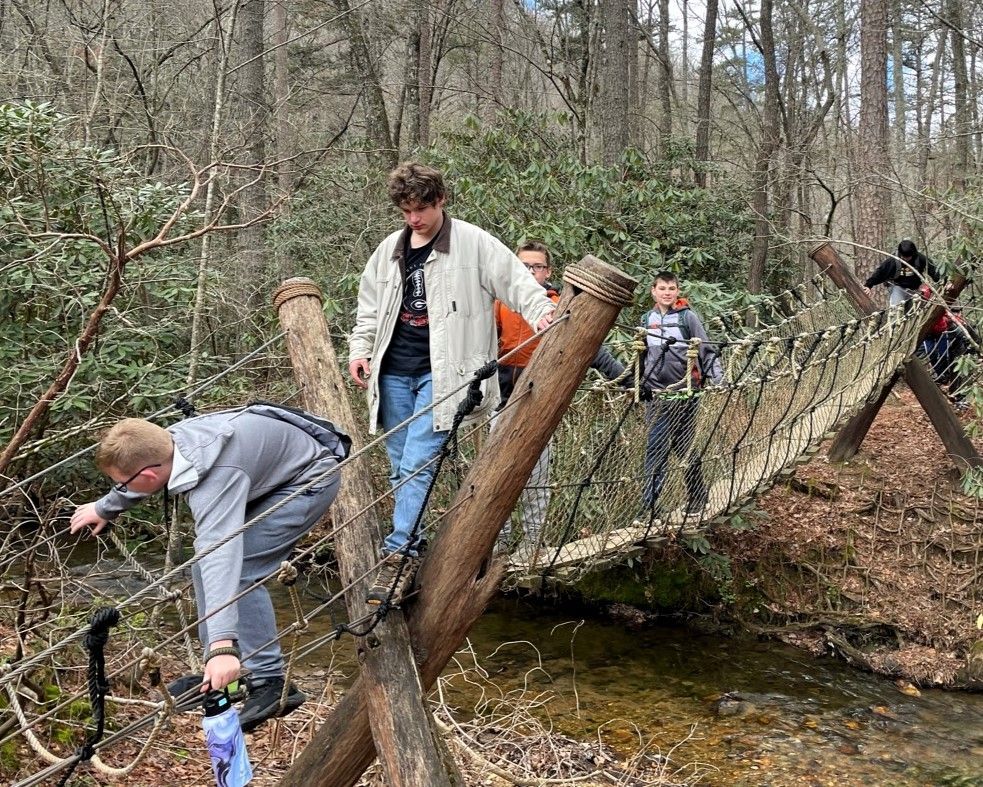 A group of people are crossing a rope bridge over a stream in the woods.