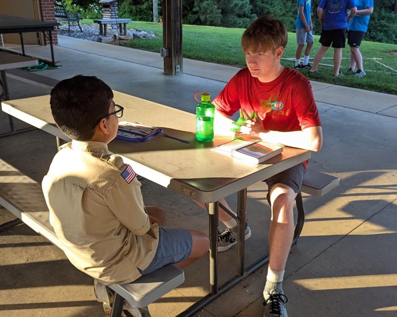 Two boys are sitting at a picnic table talking to each other.