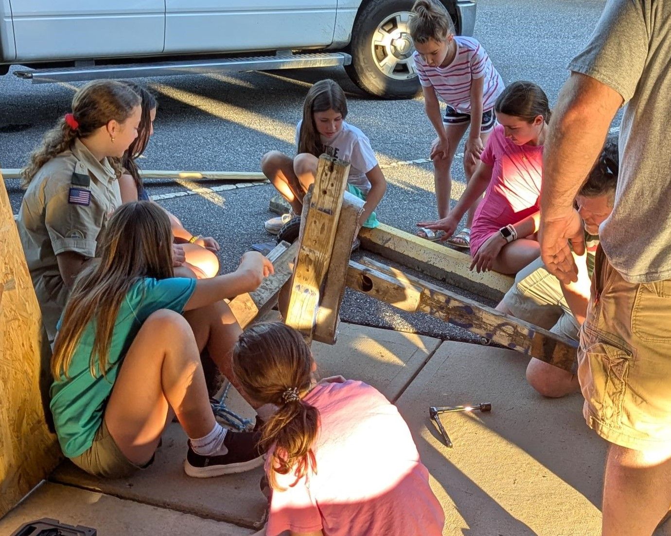 A group of young girls are sitting on the sidewalk looking at a piece of wood.